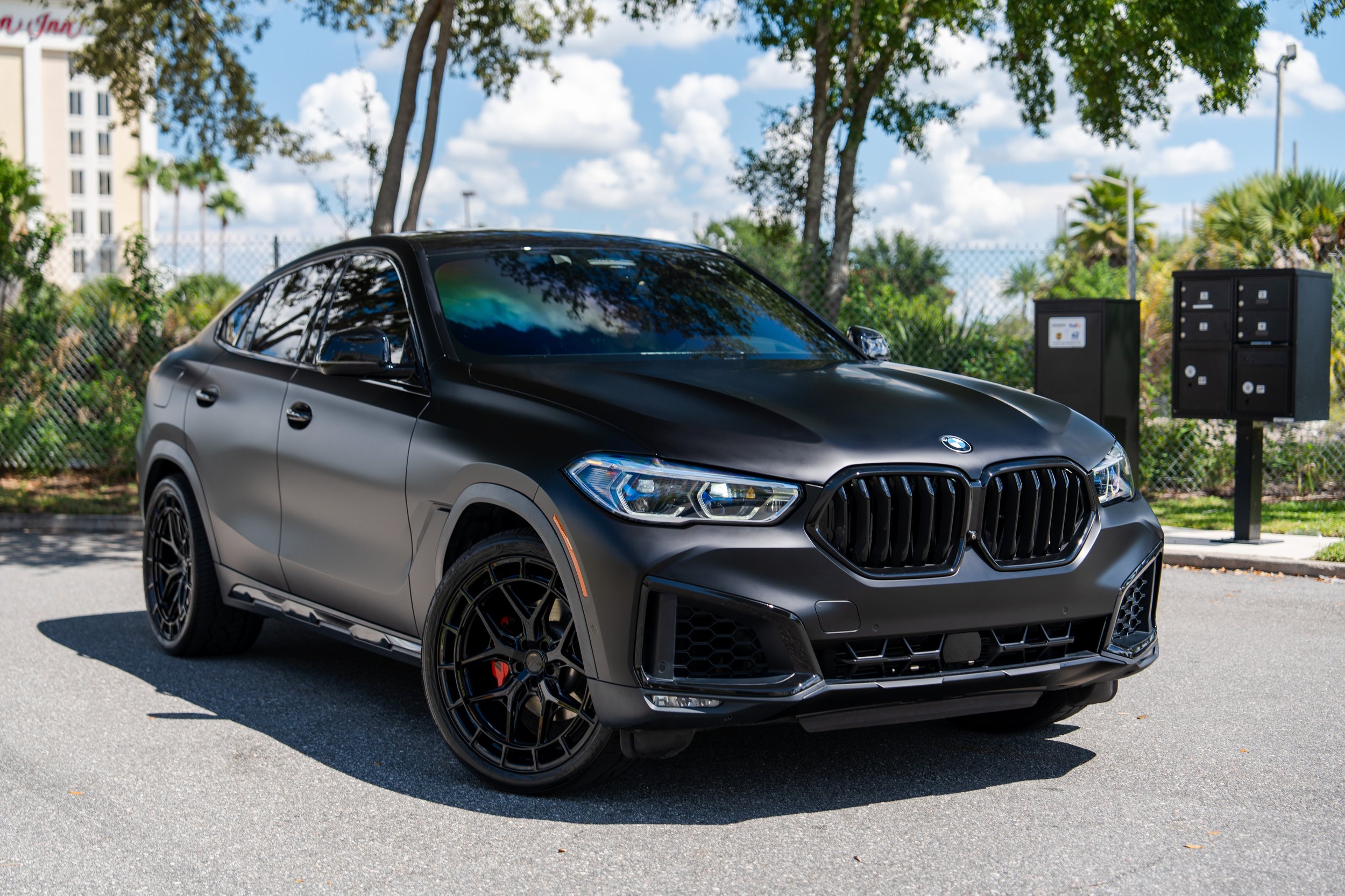 A black BMW SUV parked outdoors on a sunny day with trees, fence, and mailboxes in the background.