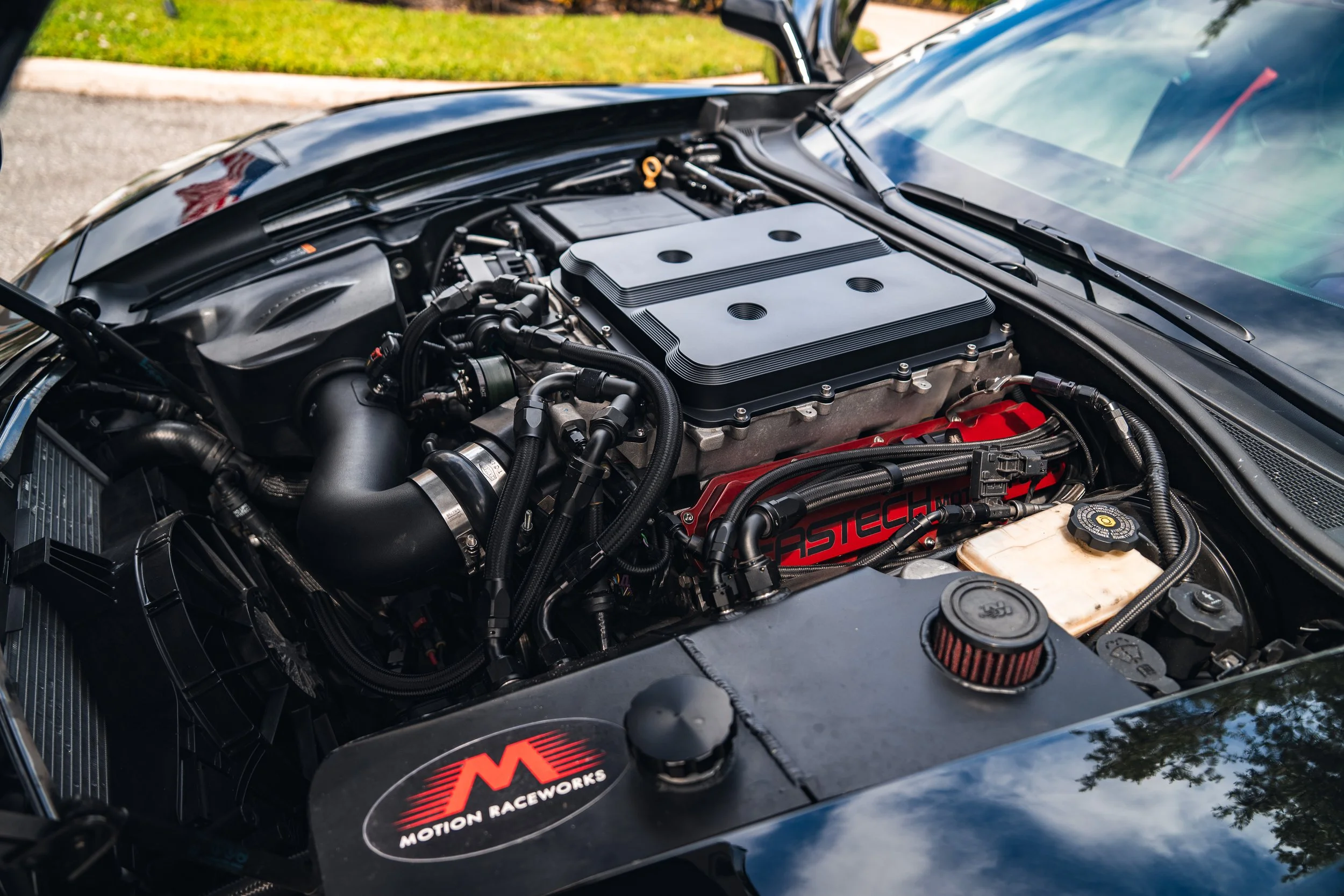 Close-up of a car engine with black cover, red lettering, and various hoses and components, including a Motions Raceworks sticker on the black panel in the front.