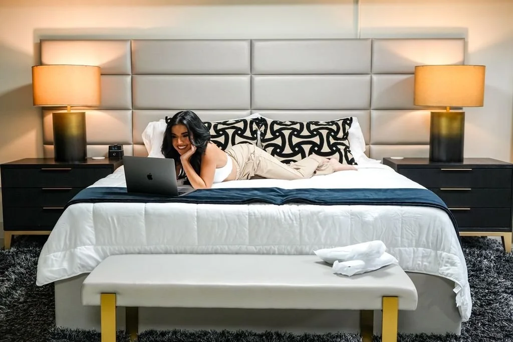A woman with dark hair lying on her stomach on a bed, using a laptop, in a modern hotel room with symmetrical nightstands and lamps on each side of the bed.
