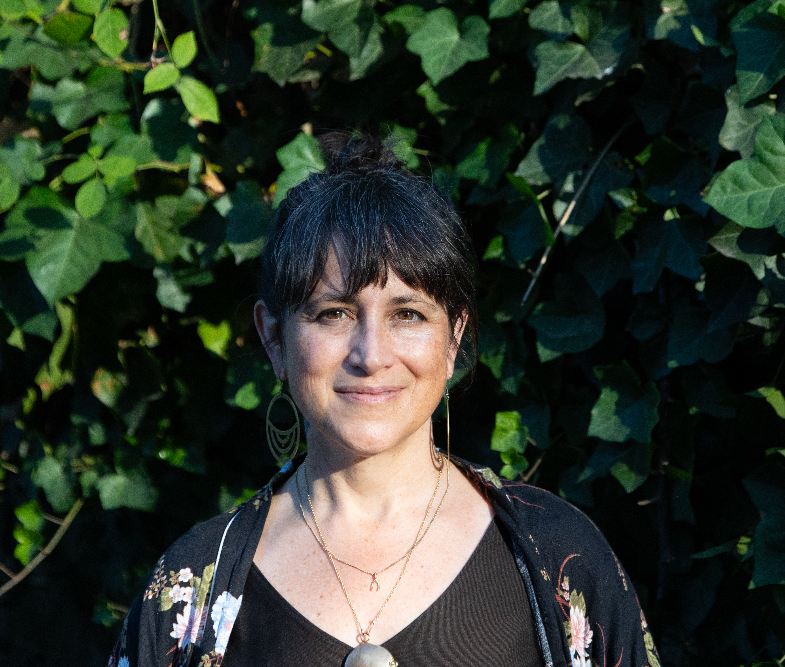A white woman with brown hair and eyes stands in front of green ivy. She has a bun, is wearing round earrings, necklaces, and black shirts, one with flowers on it.
