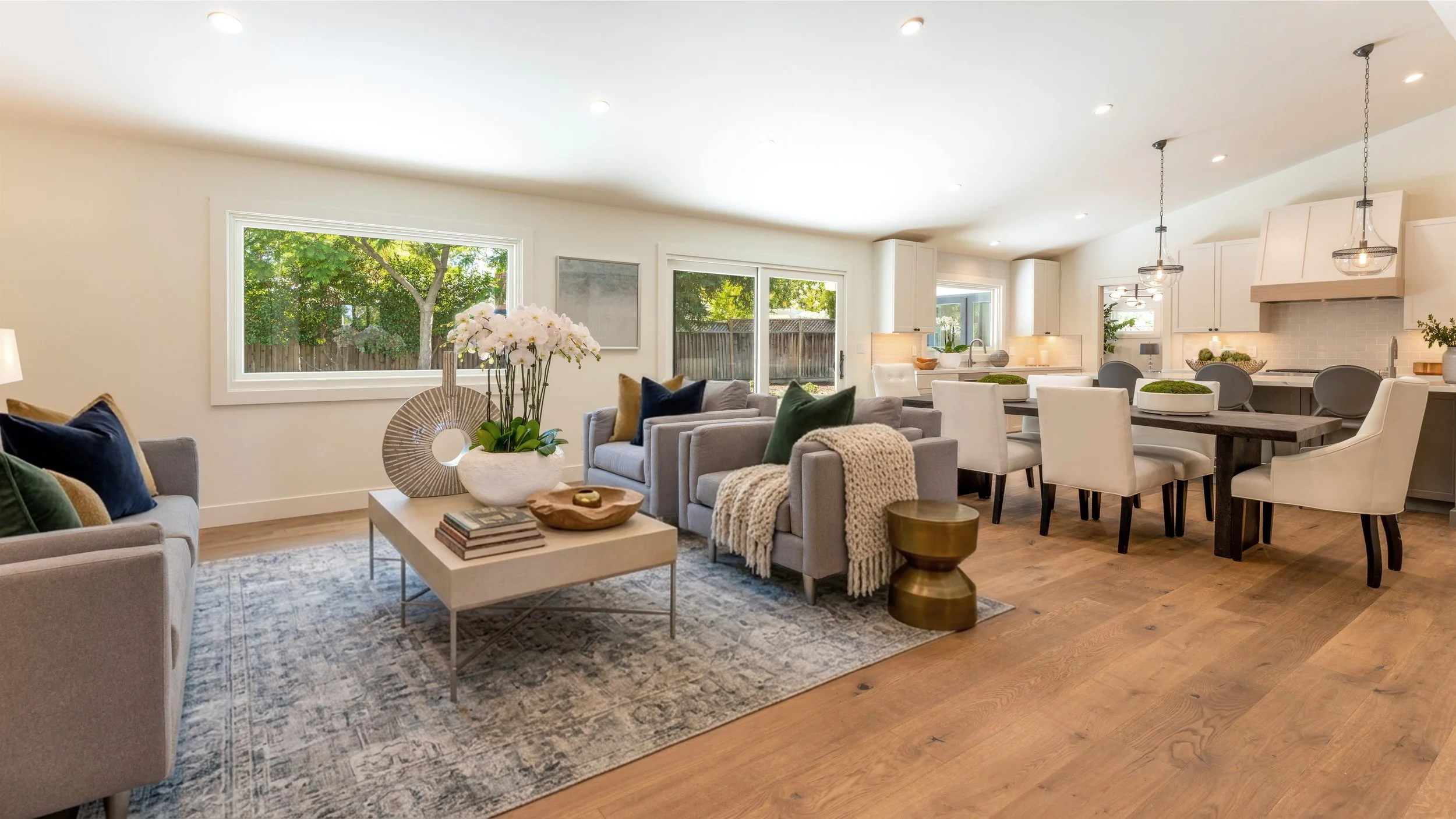 Open-concept living room and kitchen with large windows, white cabinetry, dining table, and modern decor.