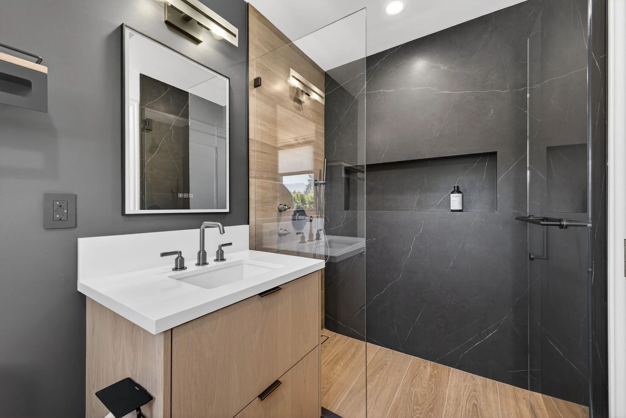 Modern bathroom with a white vanity, wooden cabinet, black and gray walls, large mirror, and walk-in shower with black marble tiles.