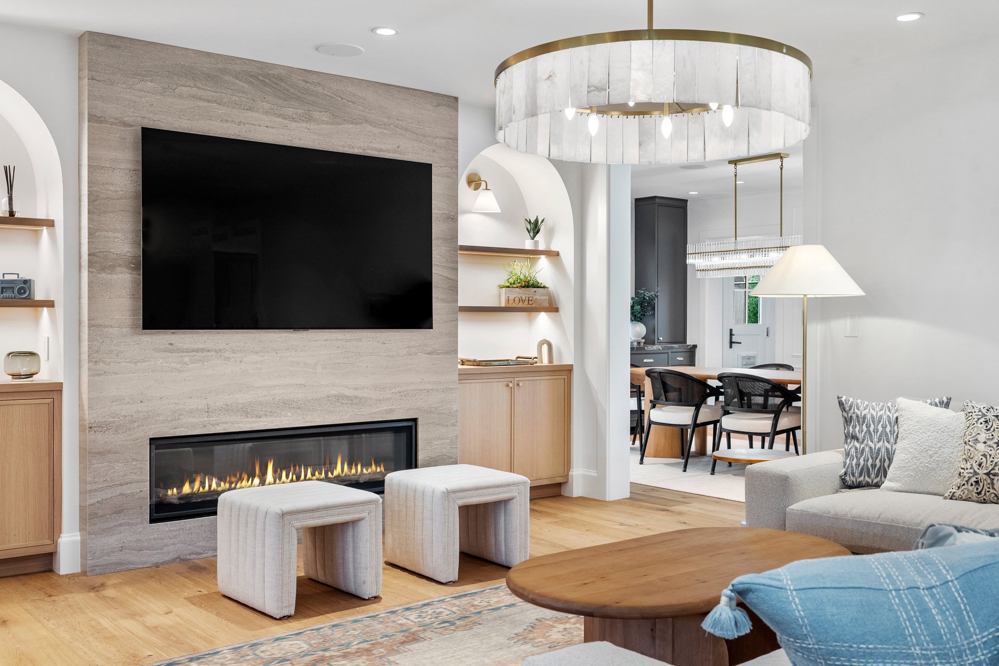 Living room with a wall-mounted TV above a fireplace, white ottomans, a chandelier, a sofa with patterned pillows, and a glimpse into the dining area with a chandelier and chairs.