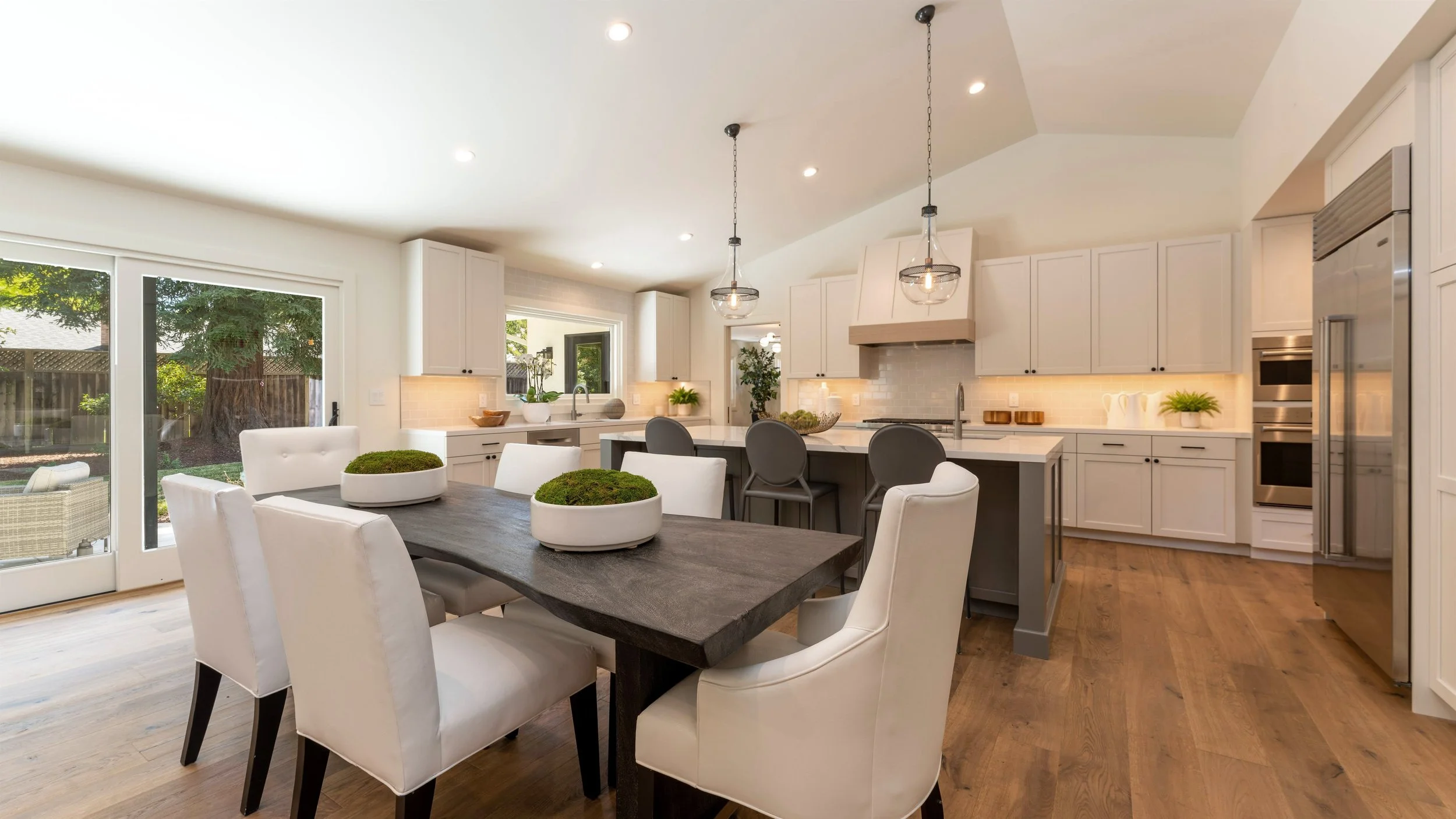 Modern kitchen with white cabinets, island, and dining table with white chairs, large window, and pendant lighting.