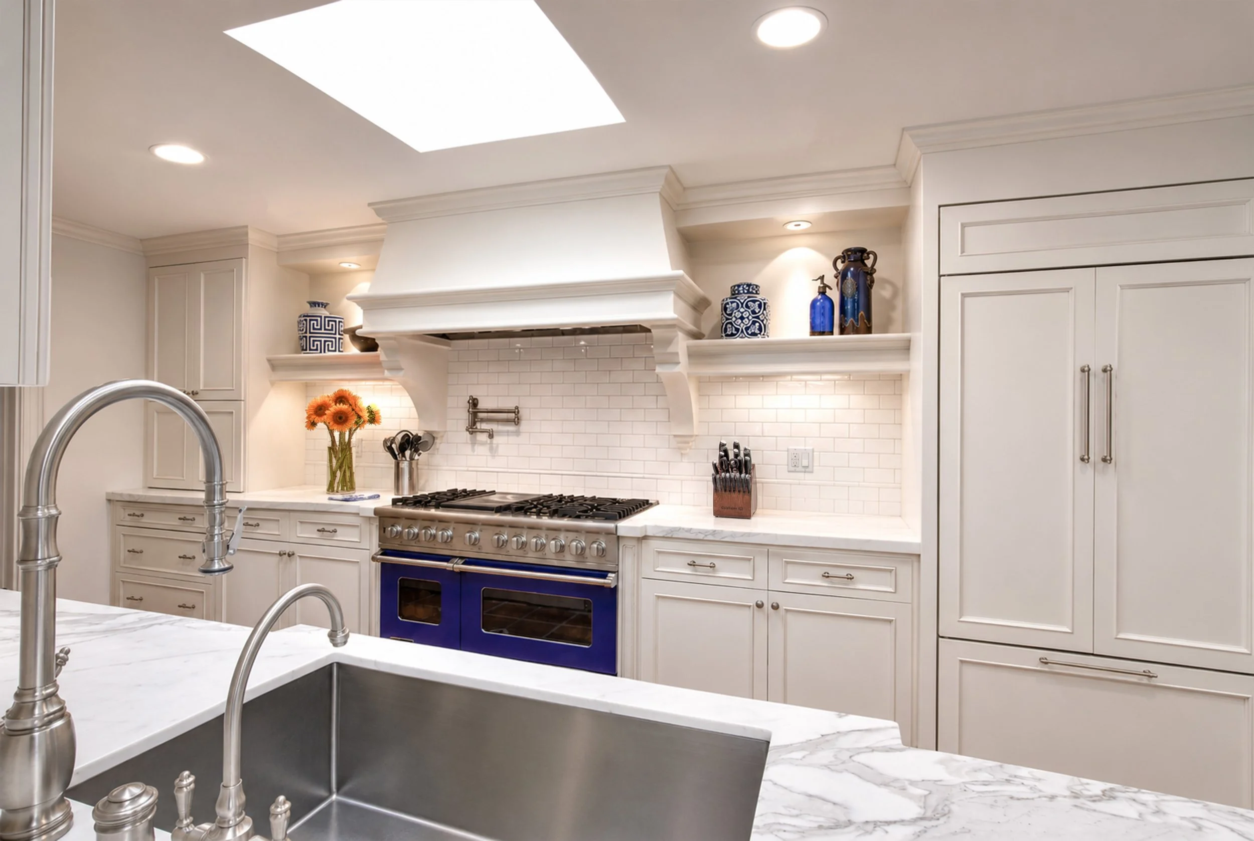 A modern kitchen with white cabinets, a purple oven, white marble countertops, a stainless steel sink, a stove, decorative vases on shelves, and a bouquet of orange flowers.