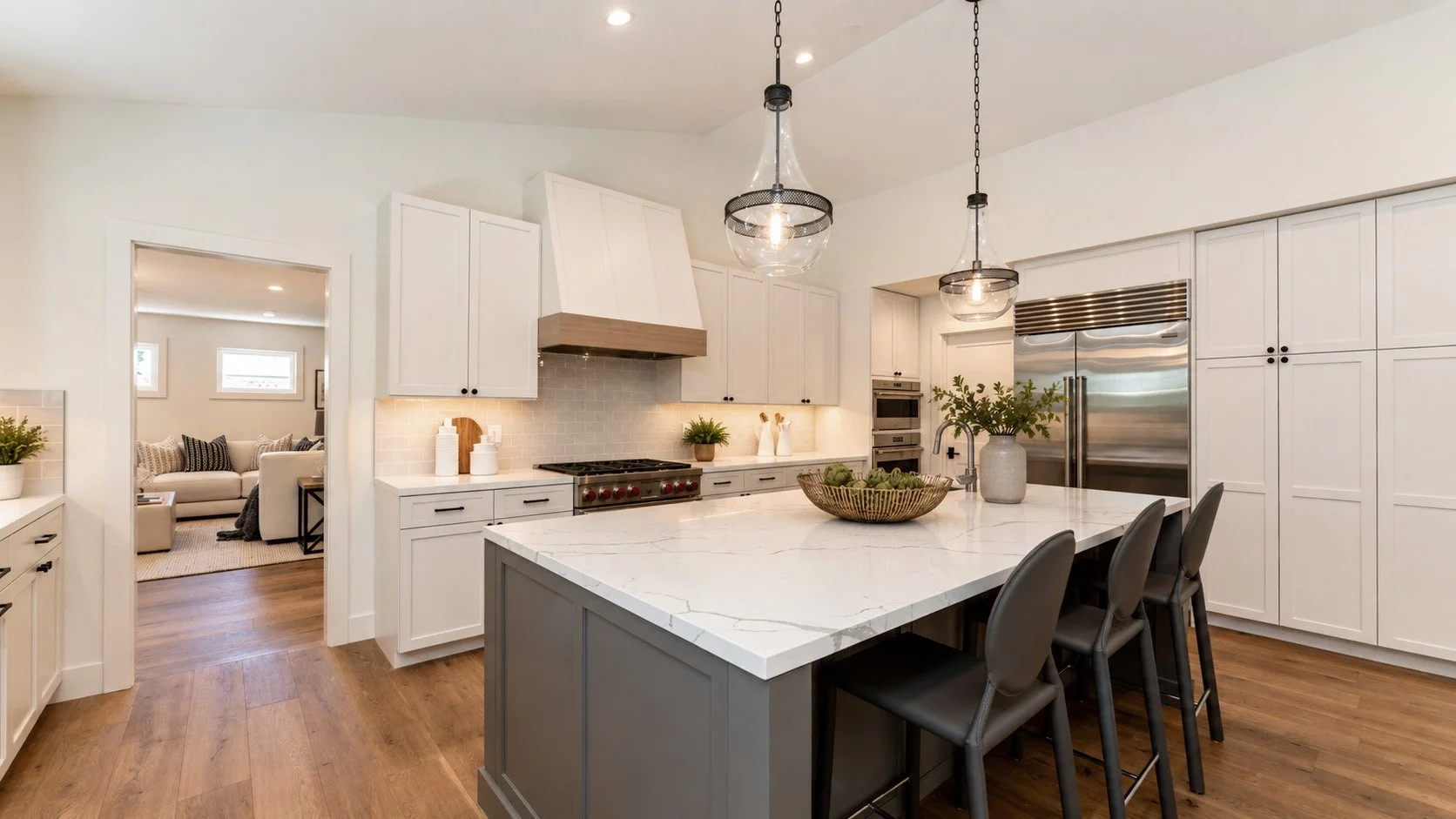 Modern kitchen with white cabinetry, a large marble island, stainless steel appliances, and wooden floors. There are pendant lights hanging from the ceiling and a view into a living room with a sofa and windows.