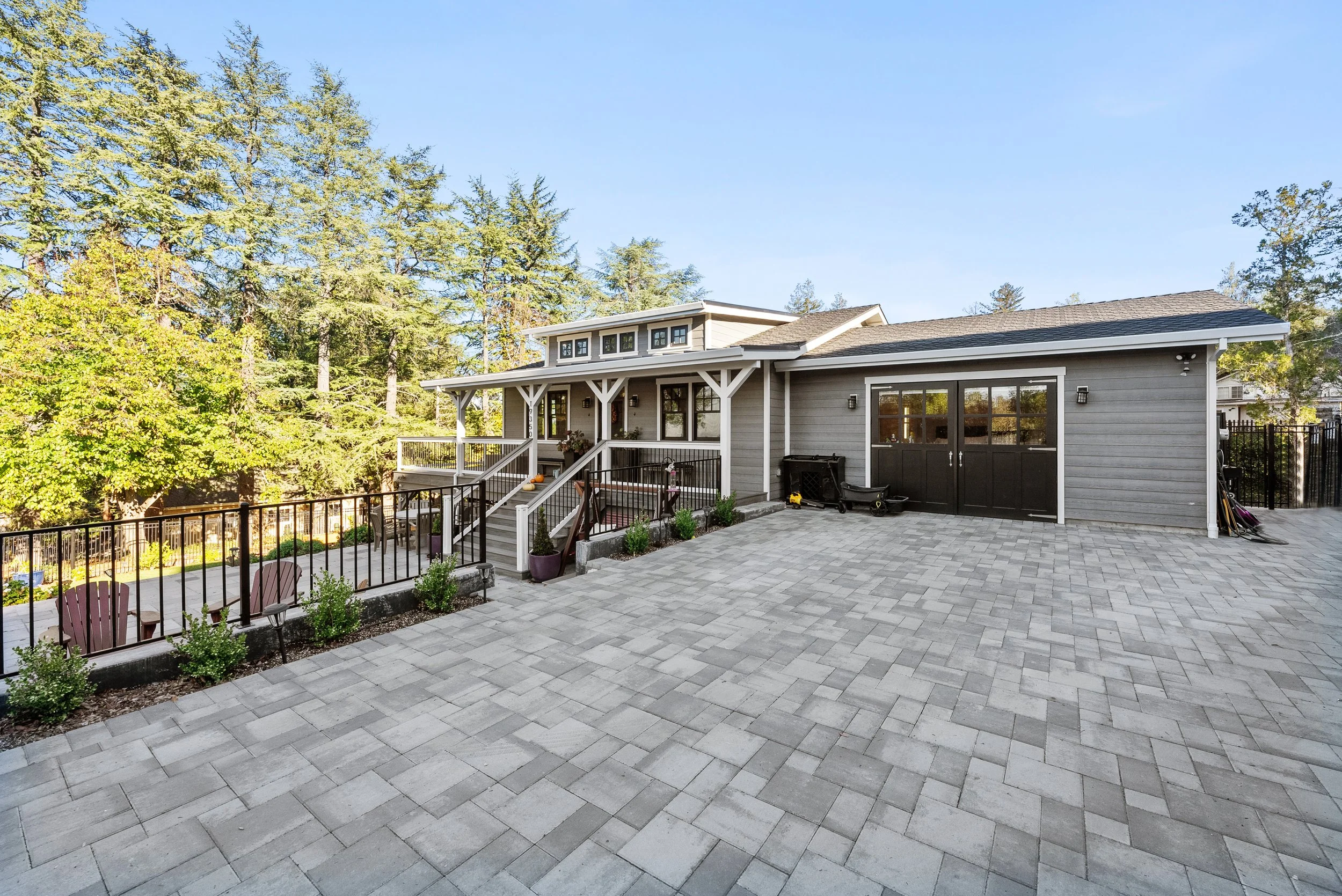 A large patio with gray paver stones in front of a gray house with a porch and black door. There are trees and a clear blue sky in the background.