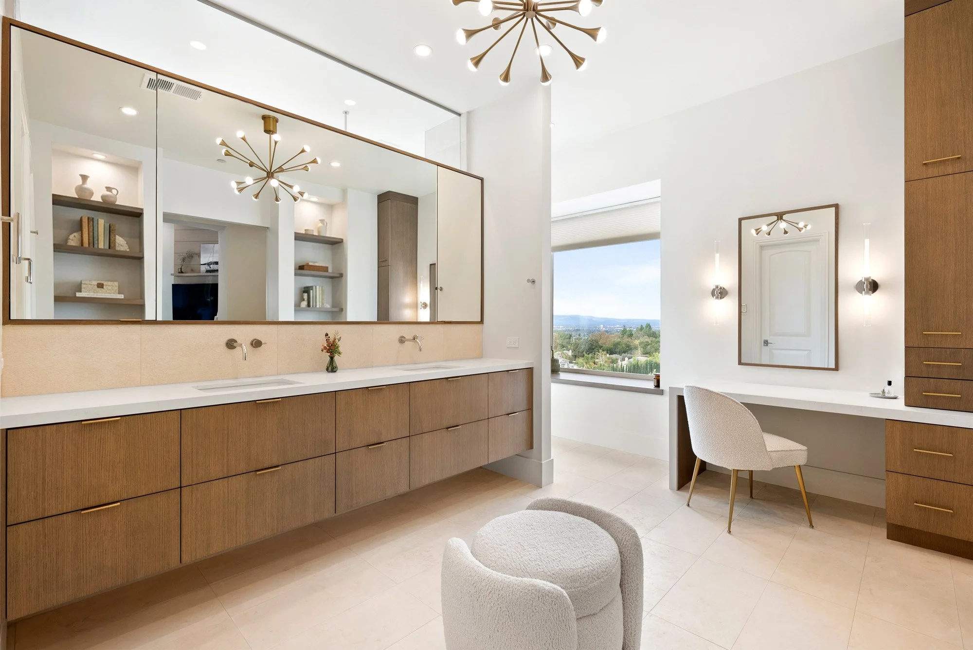 Modern bathroom vanity area featuring a large mirror, wooden cabinets, and a white countertop with two sinks. There are decorative items and books on shelves reflected in the mirror. A window offers a view of greenery and a distant landscape. The roo