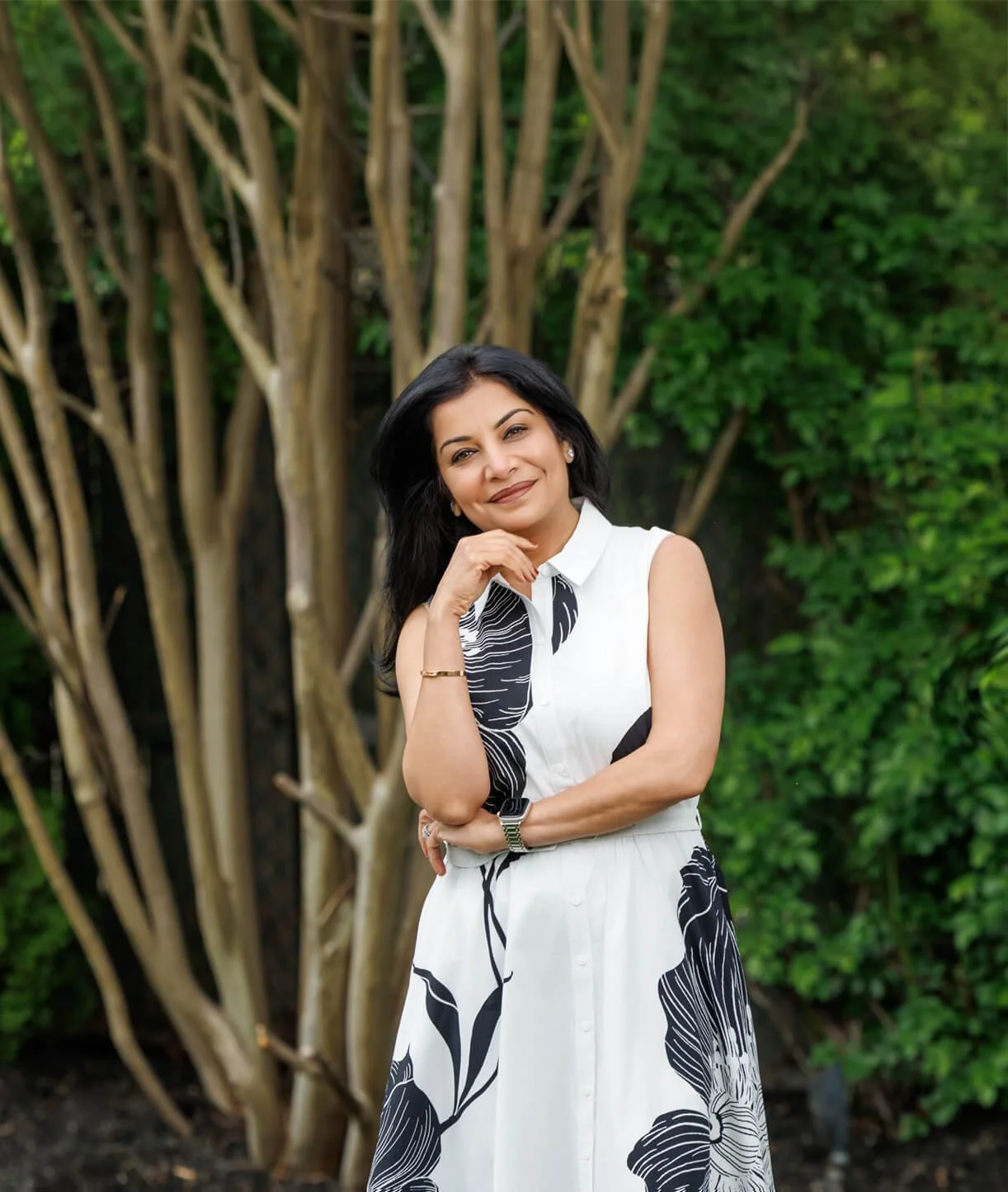Dr. Anu Gupta, a physician specializing in women's metabolic health & weight management, wearing a white top with black floral pattern, standing outdoors in front of green bushes and a tree.