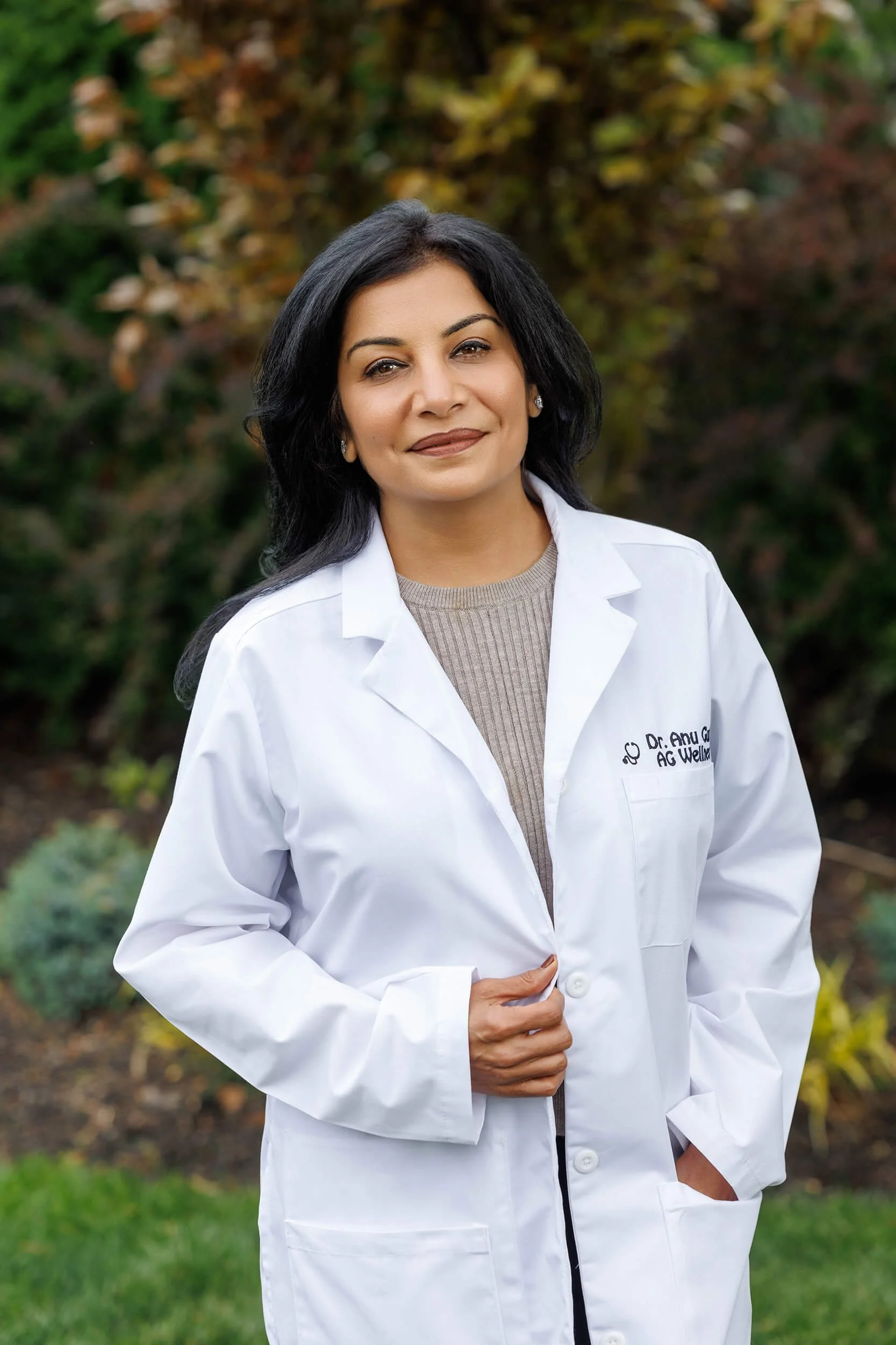 Dr. Anu Gupta, a physician specializing in women's metabolic health & weight management, wearing a white medical coat, standing outdoors with greenery and trees in the background.