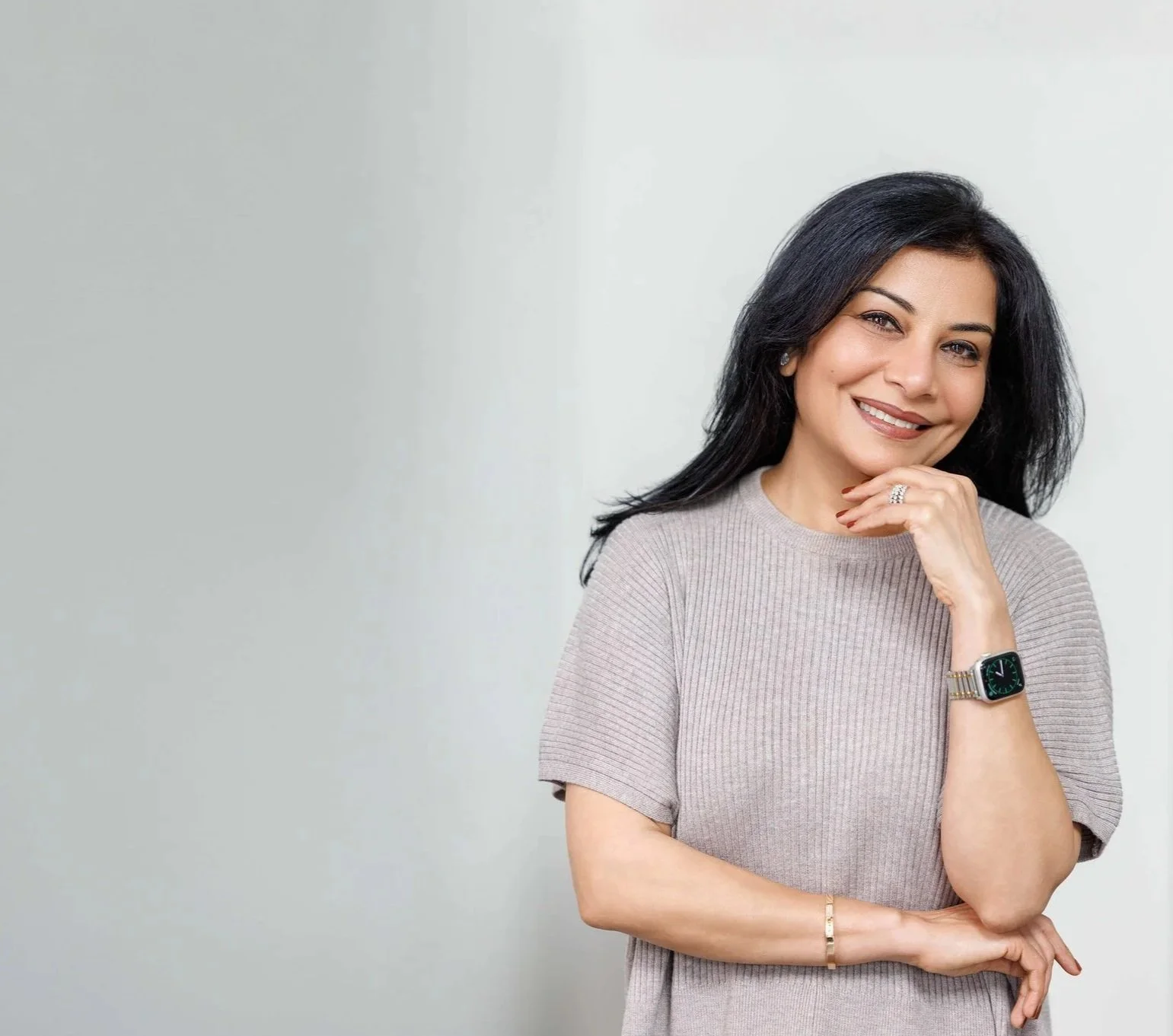 Dr. Anu Gupta, a physician specializing in women's metabolic health & weight management, wearing a beige top, a watch, and jewelry, smiling and resting her chin on her hand, standing against a plain light gray background.