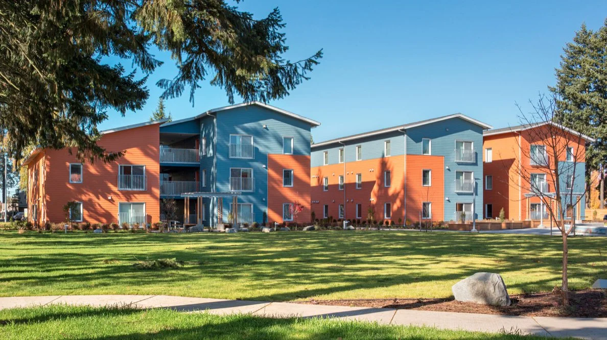 Colorful multi-story apartment building with blue and orange exterior, surrounded by a green lawn and trees under a clear blue sky.
