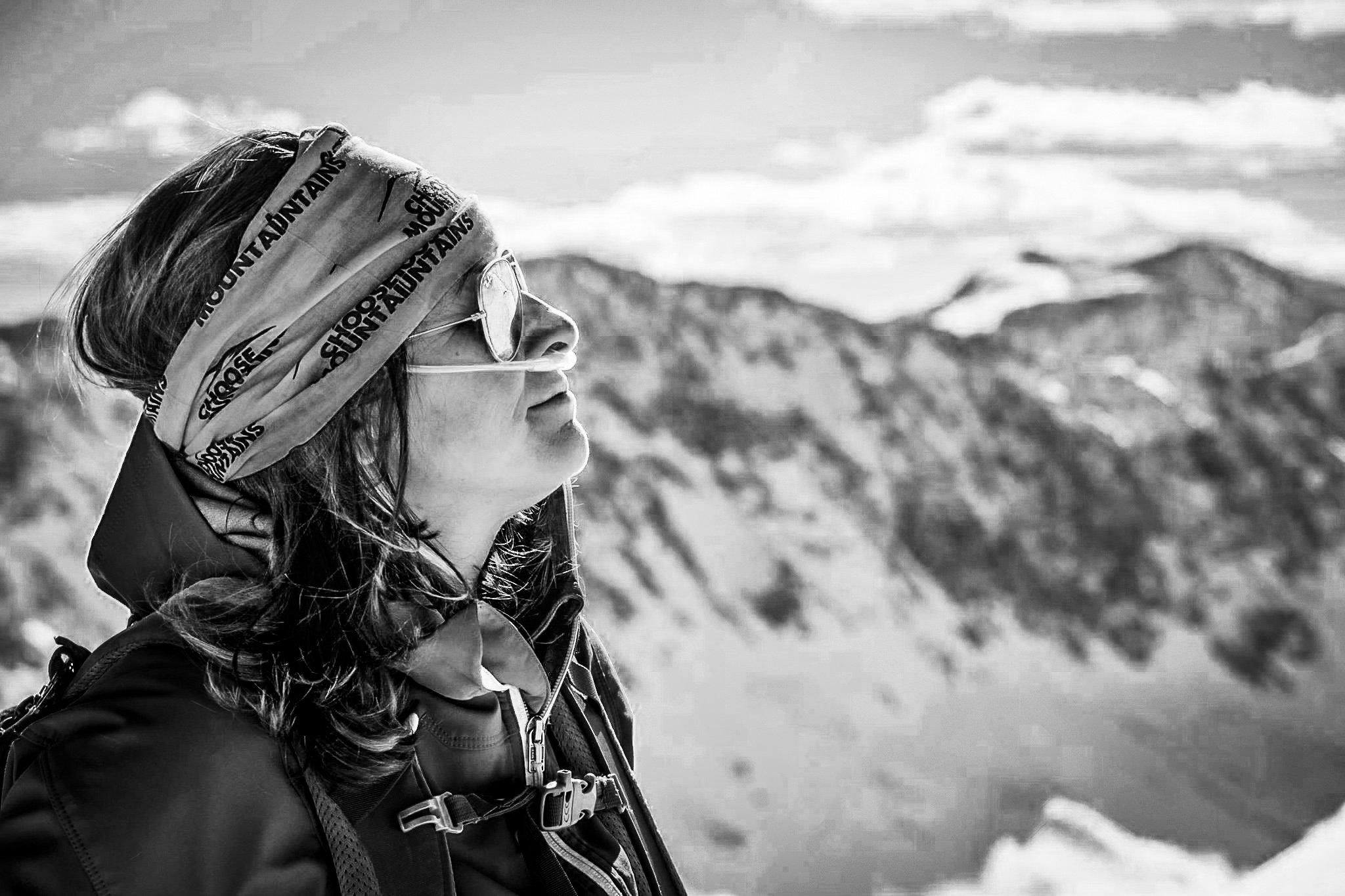 A woman with cystic fibrosis wearing sunglasses, a headband, and outdoor gear, standing in a snowy mountain landscape with mountains and sky in the background. She is hiking with supplemental oxygen and shows her oxygen nasal prongs