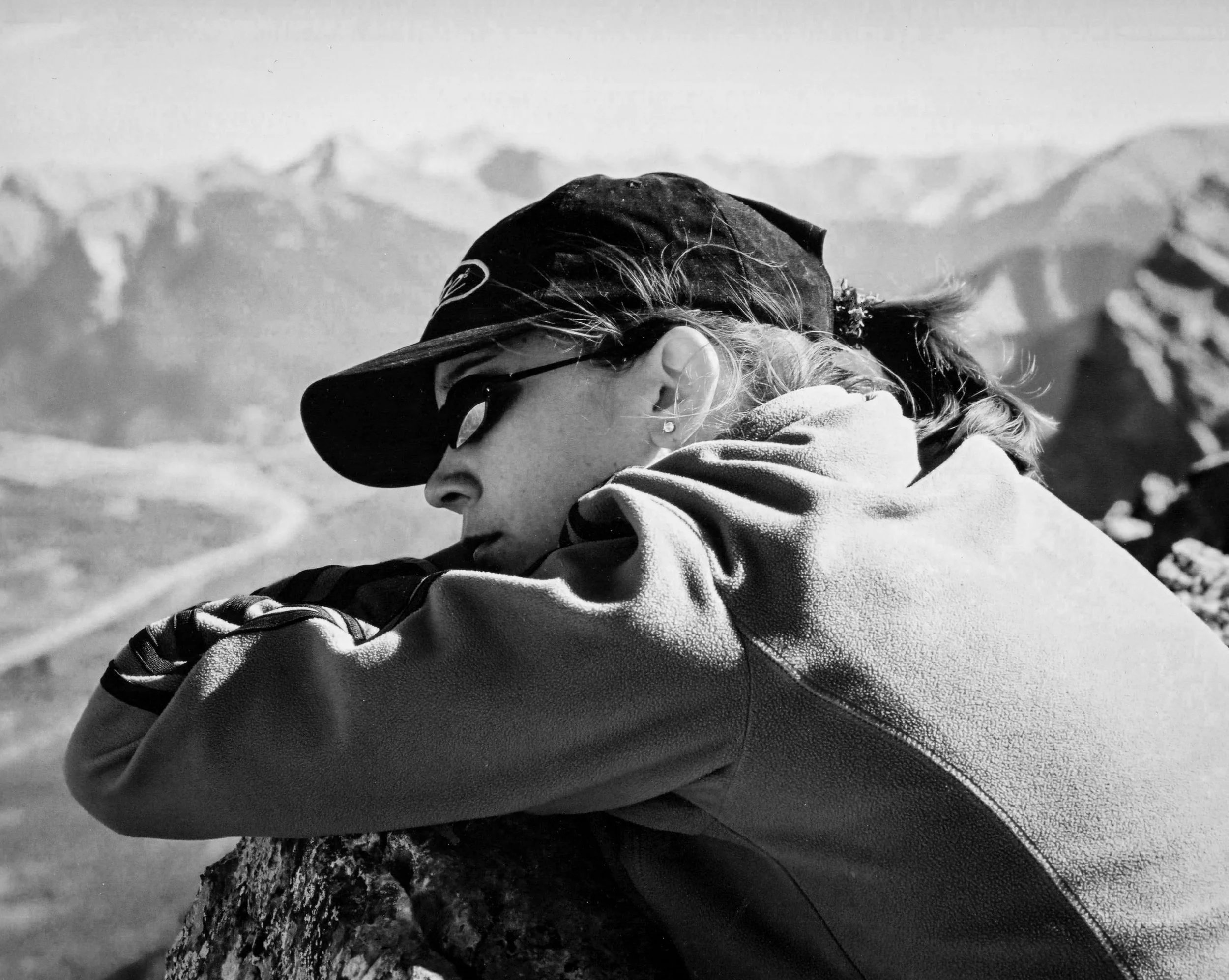 Black-and-white photo of a woman with cystic fibrosis resting her head on her arms on a rocky surface with mountain scenery in the background. She is on the summit of Ha Ling in Canmore, Alberta, Canada