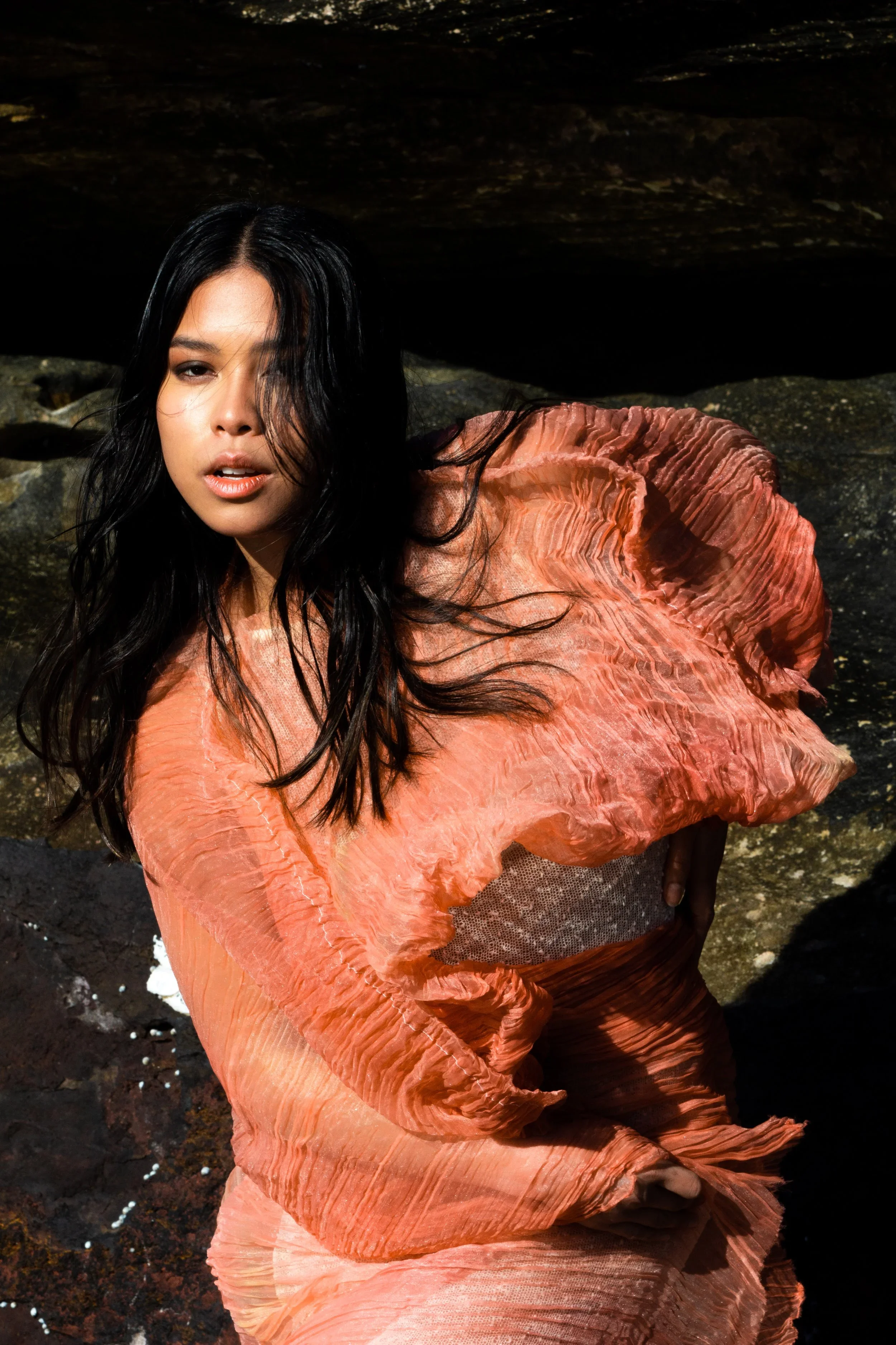 Woman with long black hair wearing a flowing coral-orange dress in front of a dark stone wall