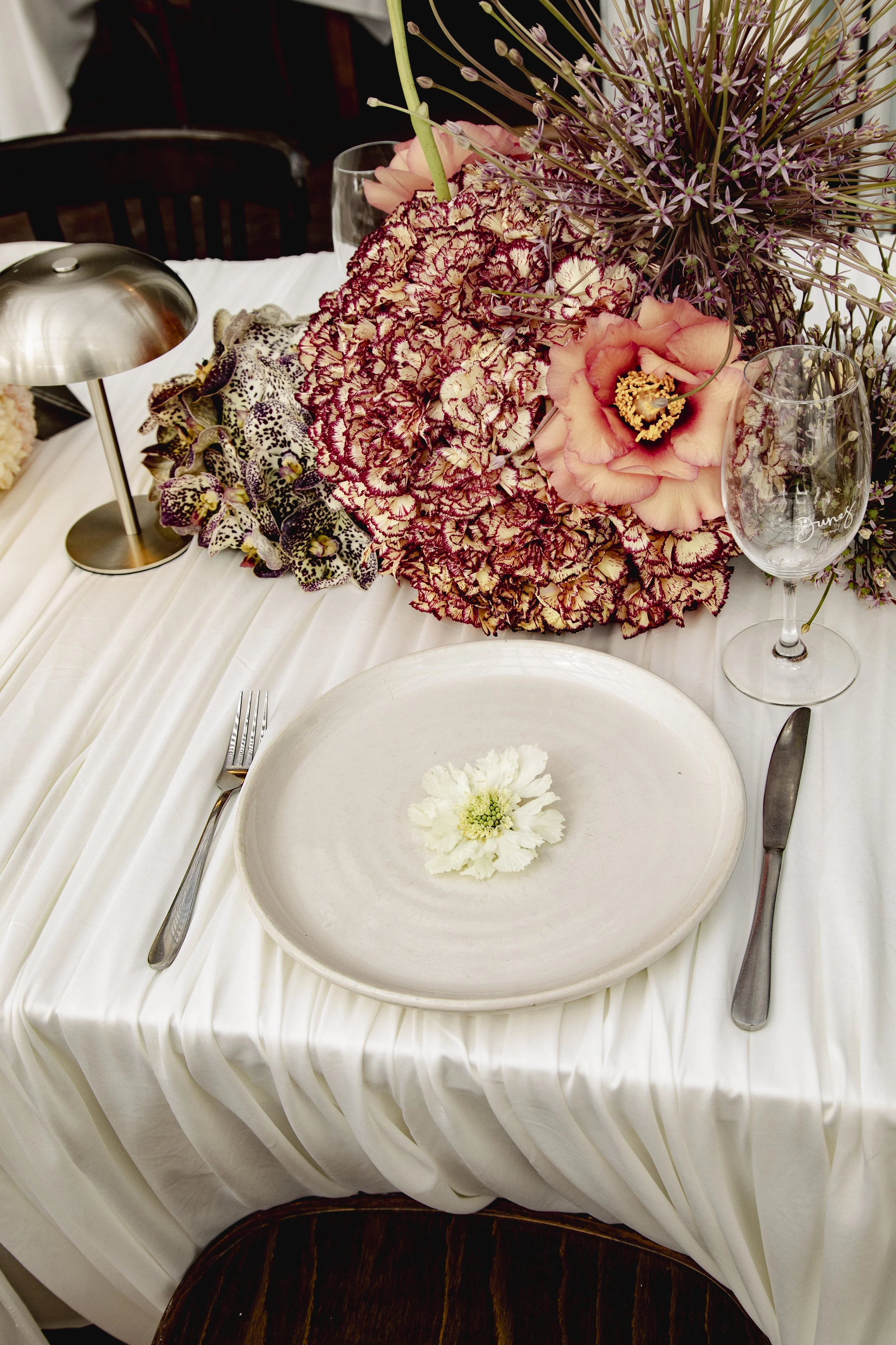 Elegant white table setting with a colorful floral centerpiece, dinner plate, silverware, wine glass, and candle lamp.