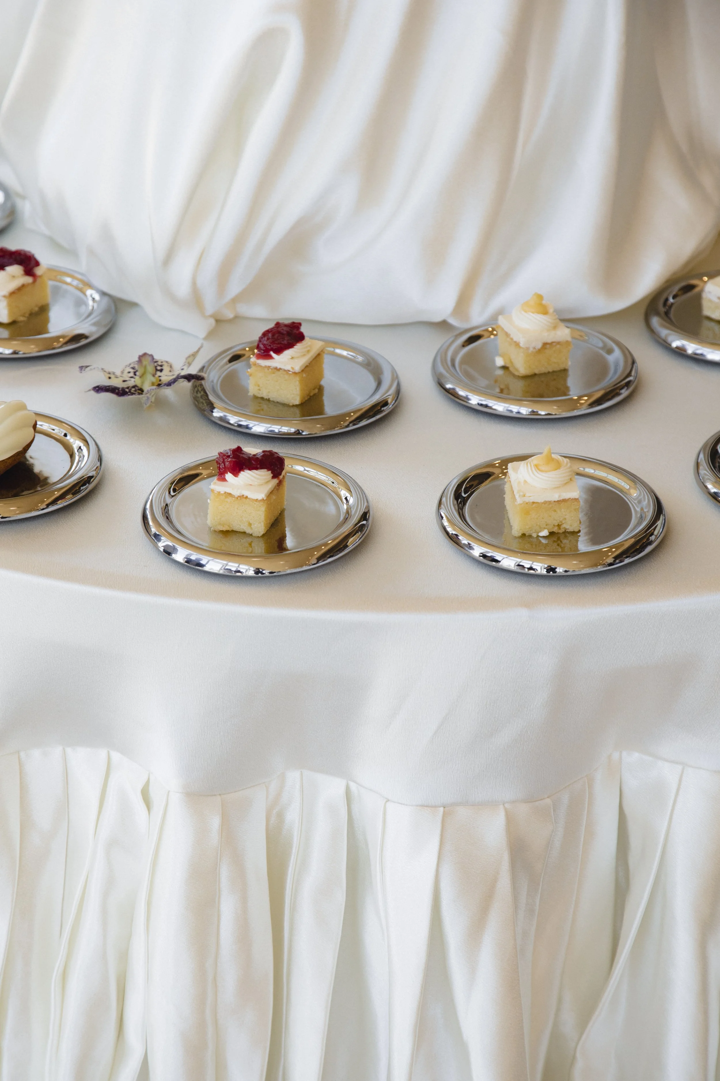 Assorted frosted cake squares on silver dessert plates arranged on a white dessert table.