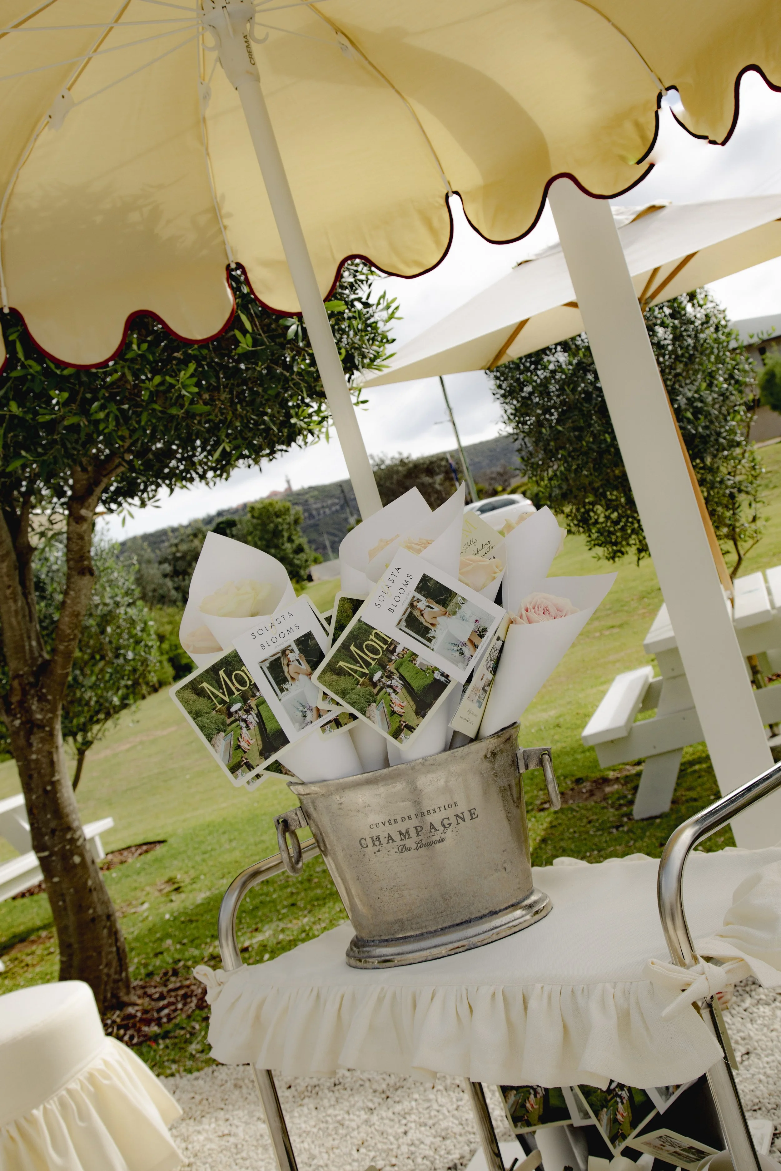 Outdoor wedding display with cream scalloped umbrellas, floral brochures, and a vintage champagne bucket in a garden