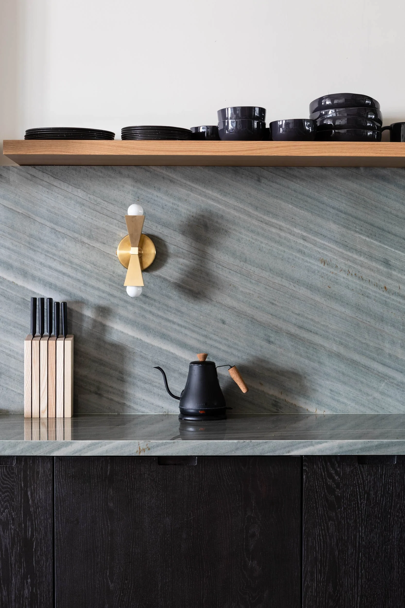 Kitchen with black teapot, wooden knife holder, black bowls and dishes on wooden shelf, modern wall sconce, grey marble wall, black cabinets.