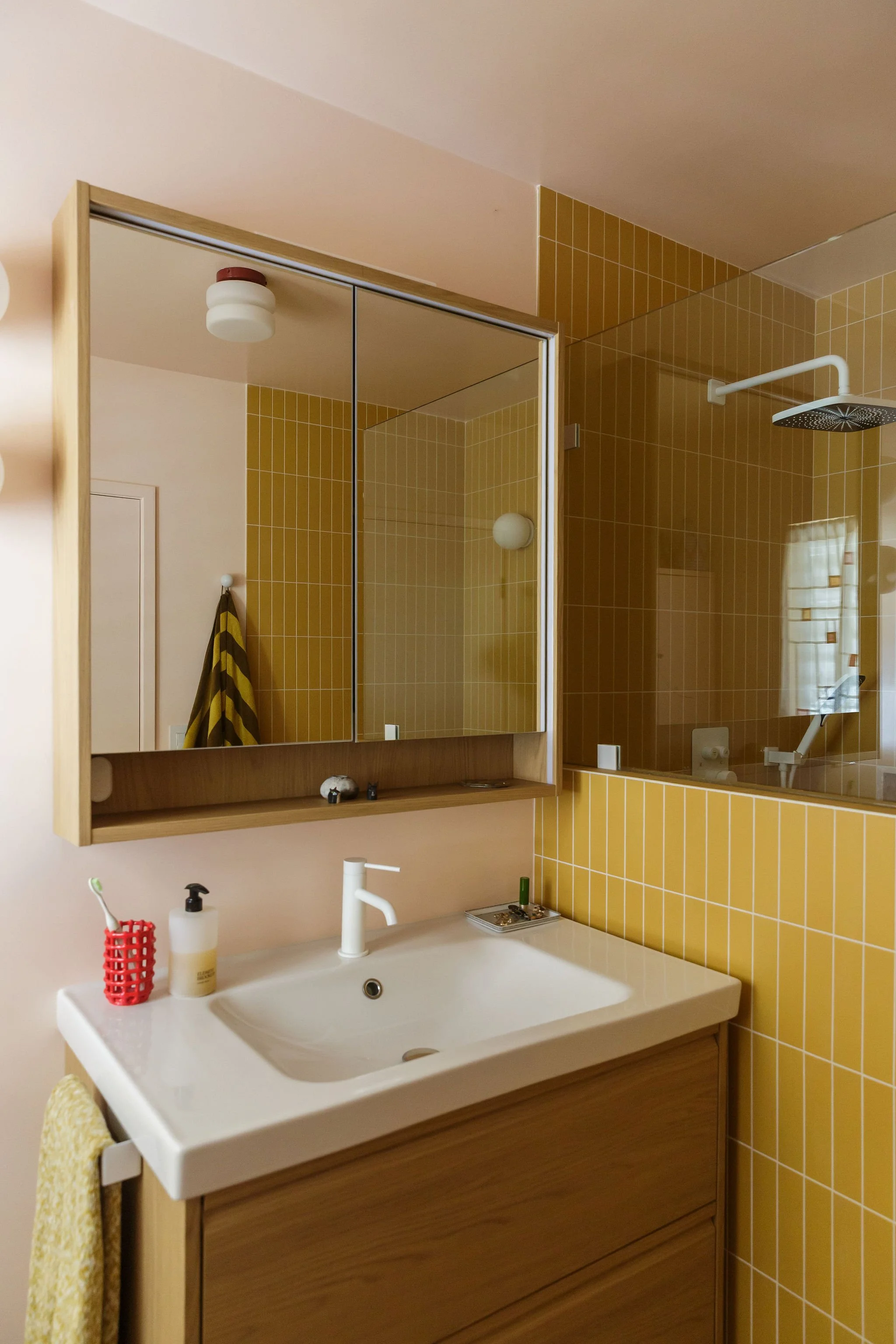 A bathroom with a white sink, wooden cabinet, mirror, yellow tiled wall, and shower area with a rainfall showerhead.