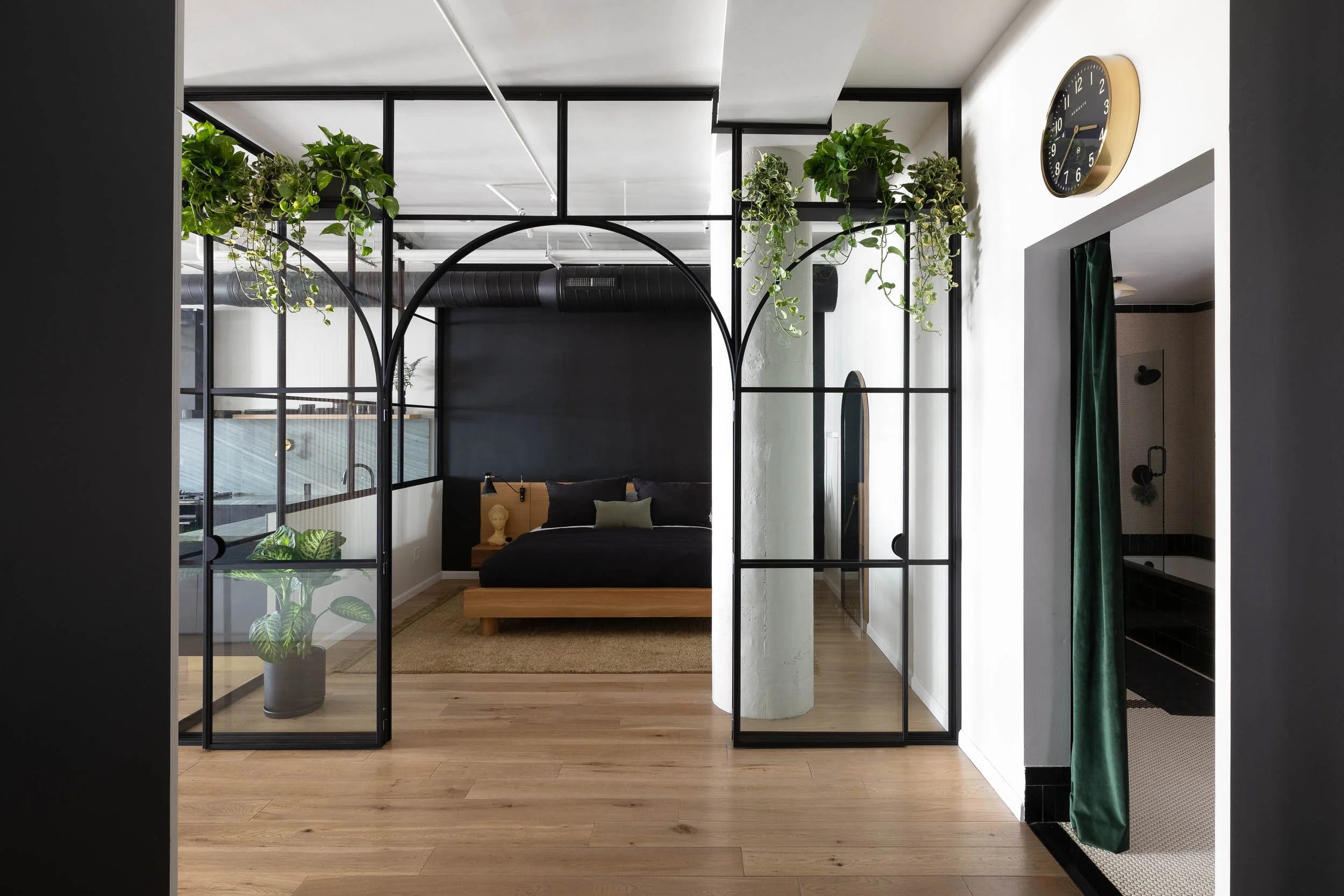 Modern bedroom with black wall, wooden bed frame, black bedding, and numerous green plants in hanging pots and on black metal frames, with nearby bathroom featuring a green curtain.