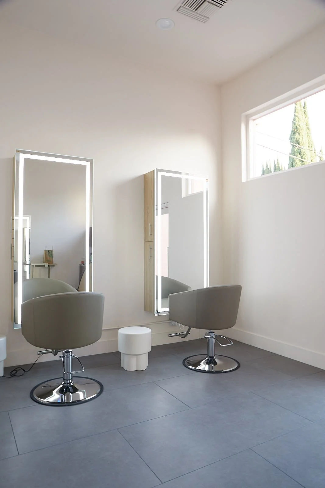 Empty hair salon with two gray styling chairs, large mirrors with lights, a white stool, and a window with sunlight.