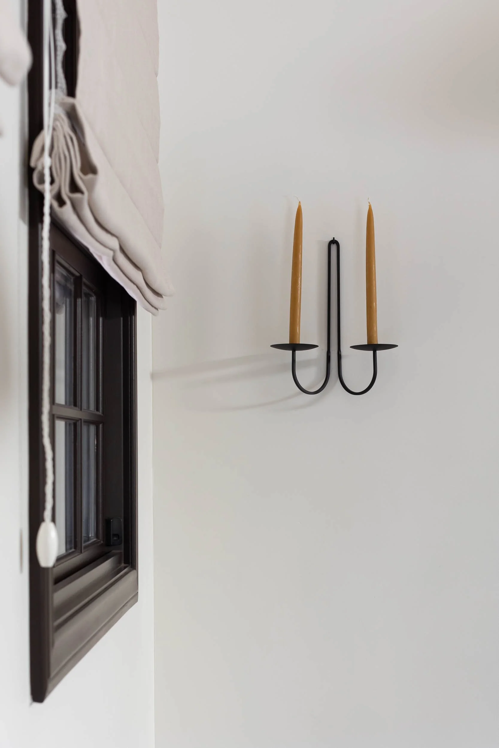 Decorative wall sconce with two lighted candles on a white wall next to a window with a beige Roman shade.