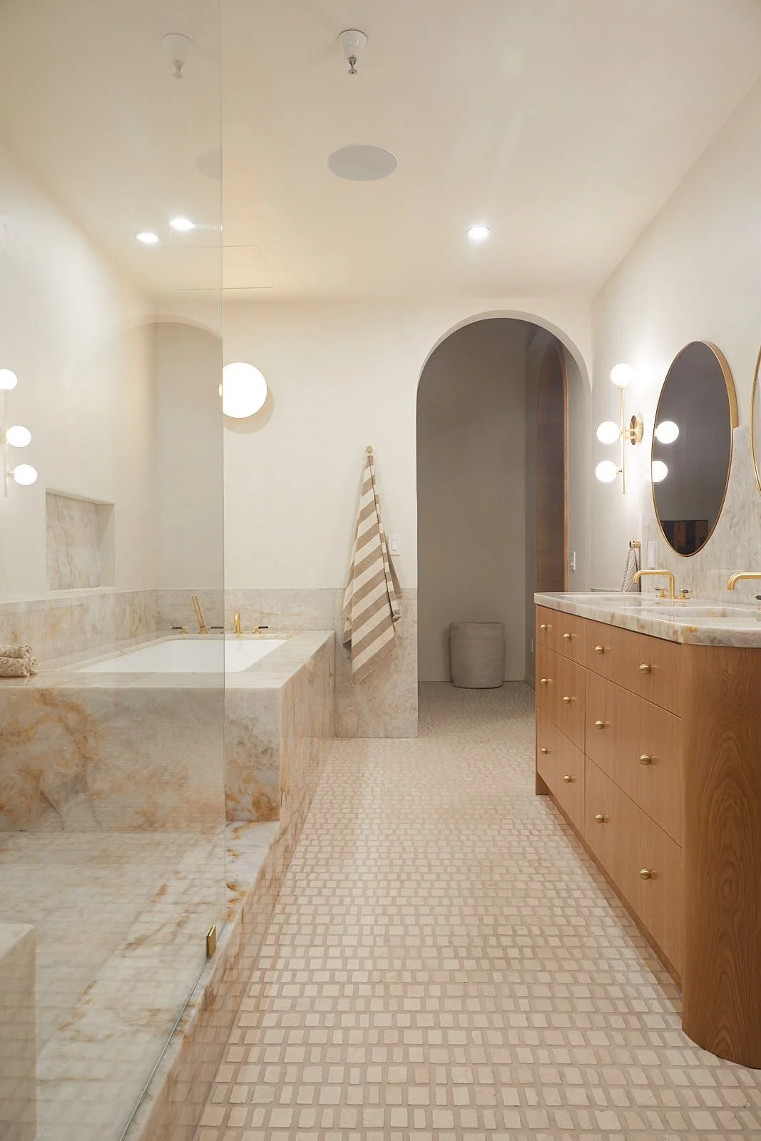 Modern bathroom with beige and brown color scheme, featuring a bathtub with marble surround, wooden vanity with marble countertop, and arched doorway.