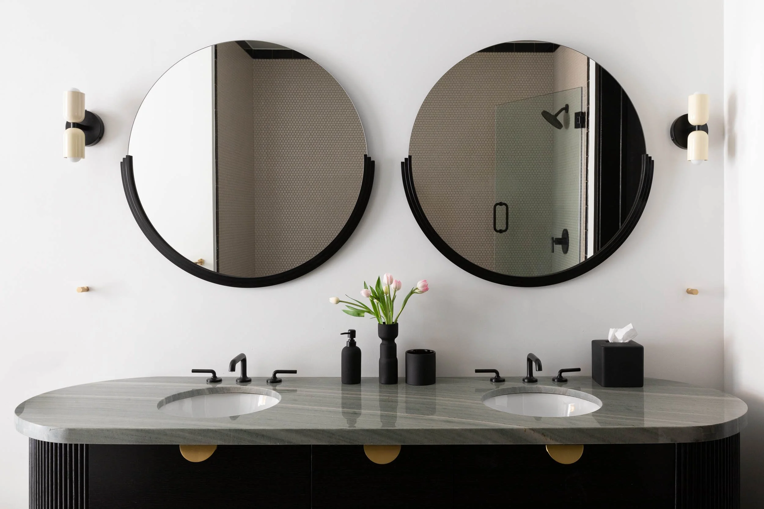 Modern bathroom double vanity with two round black-framed mirrors, black wall sconces, a marble countertop with two sinks, black faucets, a vase with pink tulips, a black soap dispenser, a black cup, and a black tissue box, with a walk-in shower visi