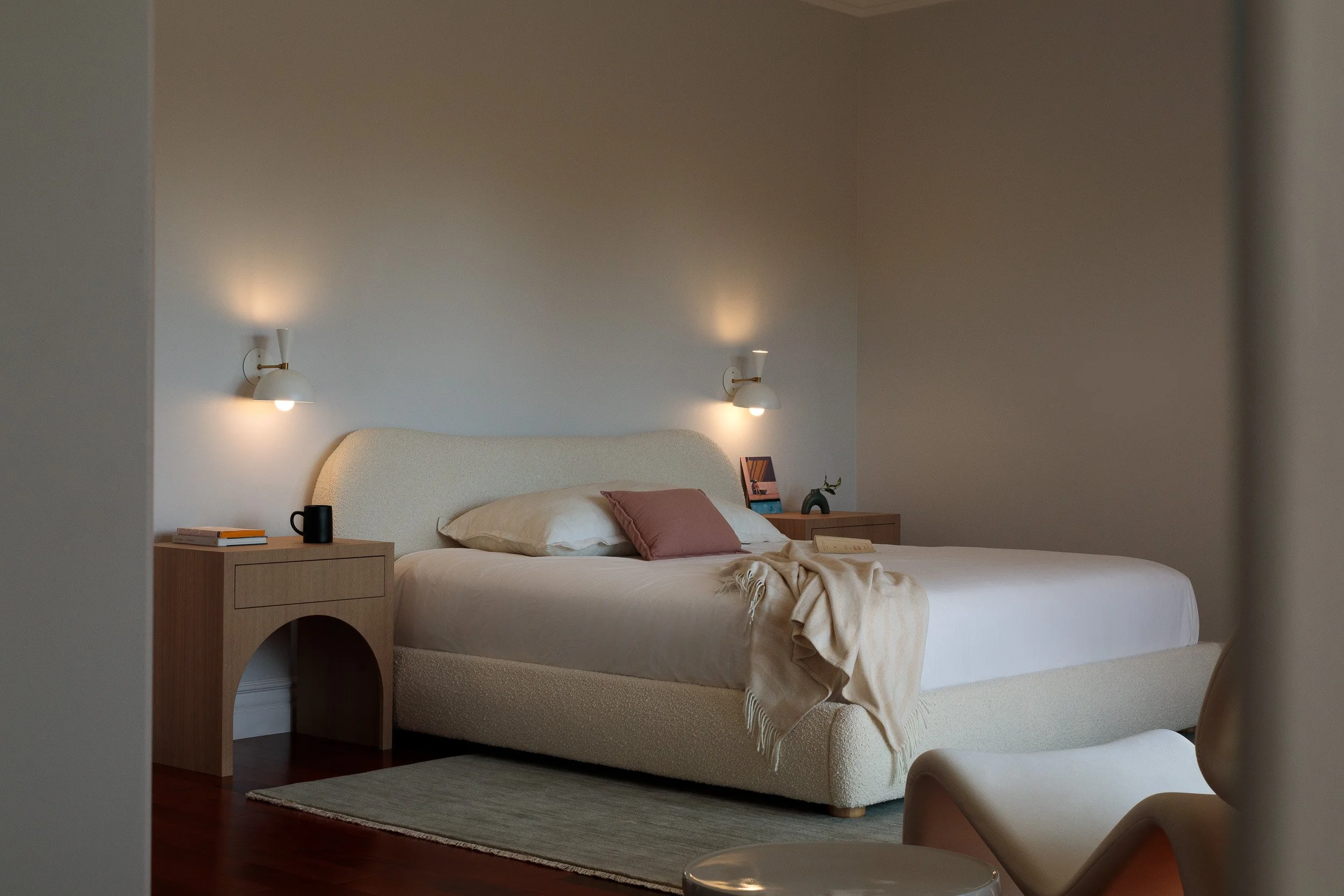 A neatly made bed with a cream-colored headboard, beige linens, and pillows, in a minimally decorated bedroom with two wall-mounted lamps, wooden nightstands with books, a black mug, and a small decorative plant.