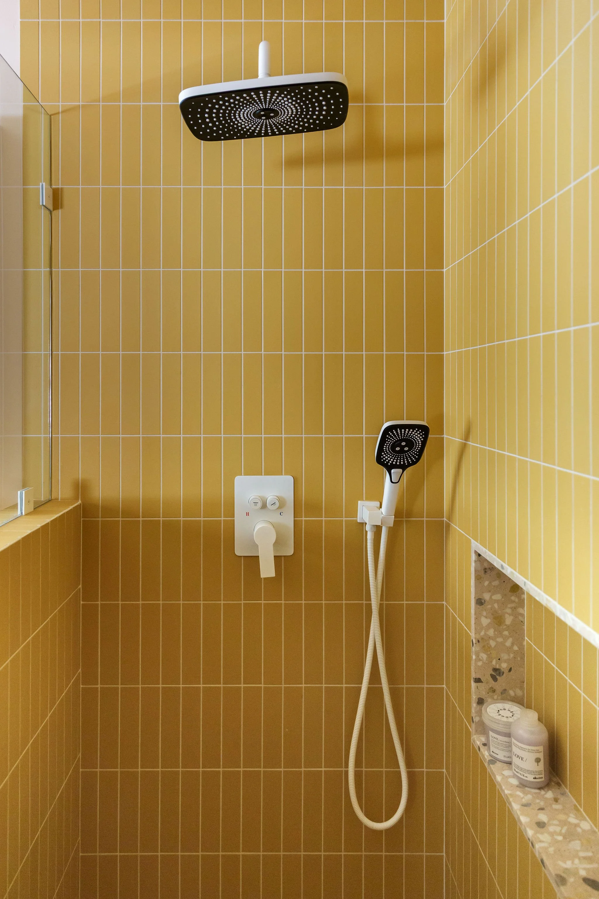 Yellow tiled shower with a rainfall showerhead, a handheld shower wand, a temperature control panel, and bottles of shower products on a tiled shelf.