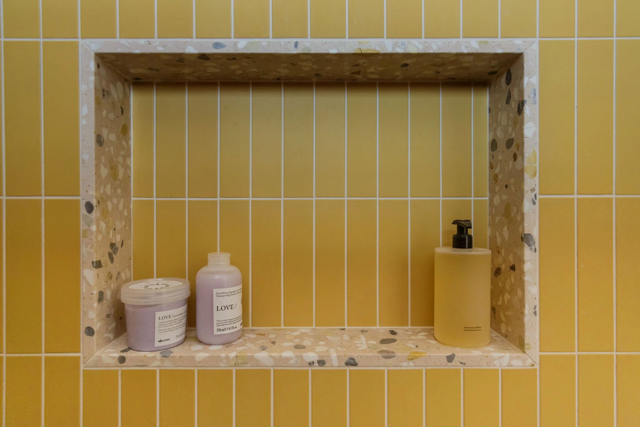 Bathroom shower niche with yellow tiles, containing hair care products.