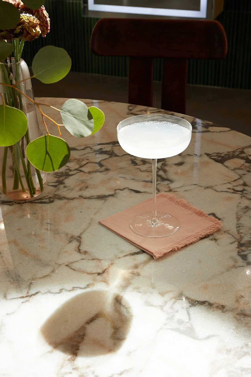 A cocktail glass filled with a white drink on a pink napkin on a marble table, with a vase of green leaves and flowers in the background.