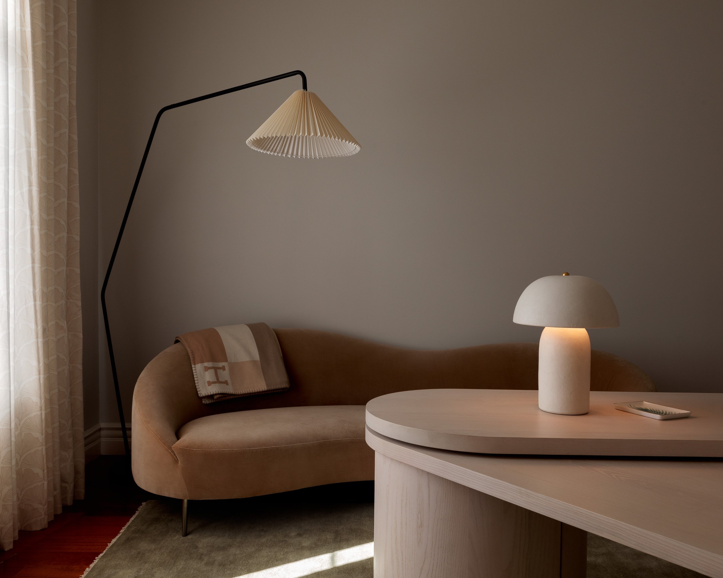 A minimalist living room corner with a beige sofa, a side table with a modern lamp, an arc floor lamp with a pleated shade, and cream curtains filtering natural light.