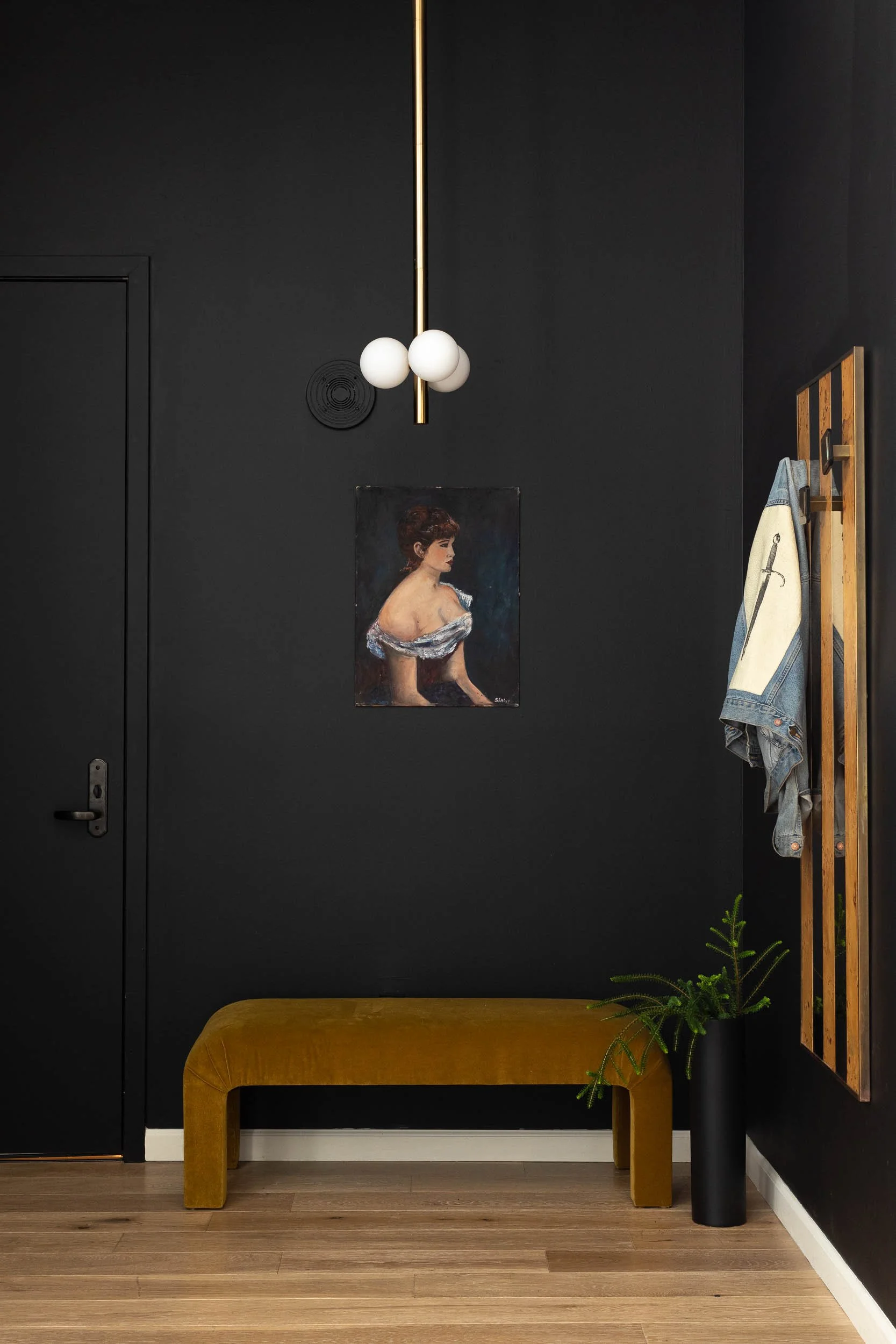 Entryway with black walls, a gold bench, a black vase with a green plant, a painting of a woman, a coat rack with a denim jacket, and a modern light fixture.