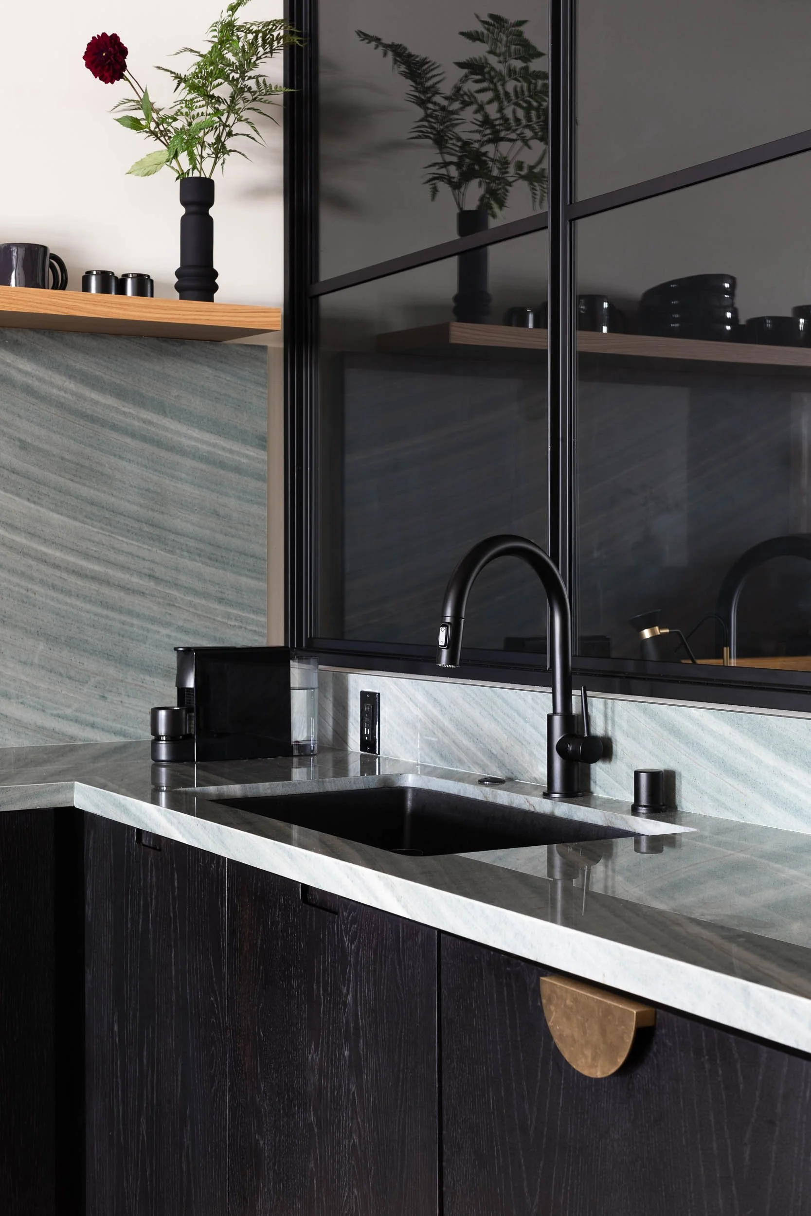 Kitchen with black faucet, black cabinetry, gray marble countertop, and a black framed window. A shelf with black dishes and a black vase with red and green flowers is visible.