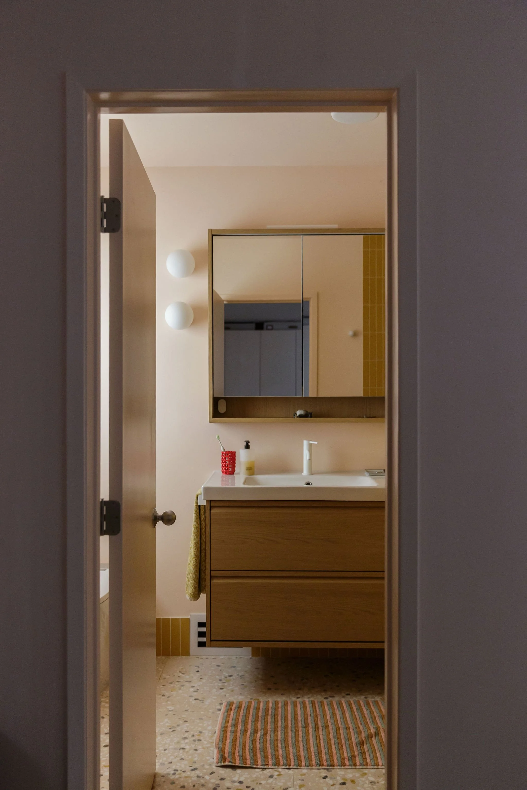 A view through a doorway into a bathroom with yellow tiled walls, a wooden vanity with a white sink, a mirror, and a striped rug on a terrazzo floor. There are white round wall lights and a soap dispenser on the sink.