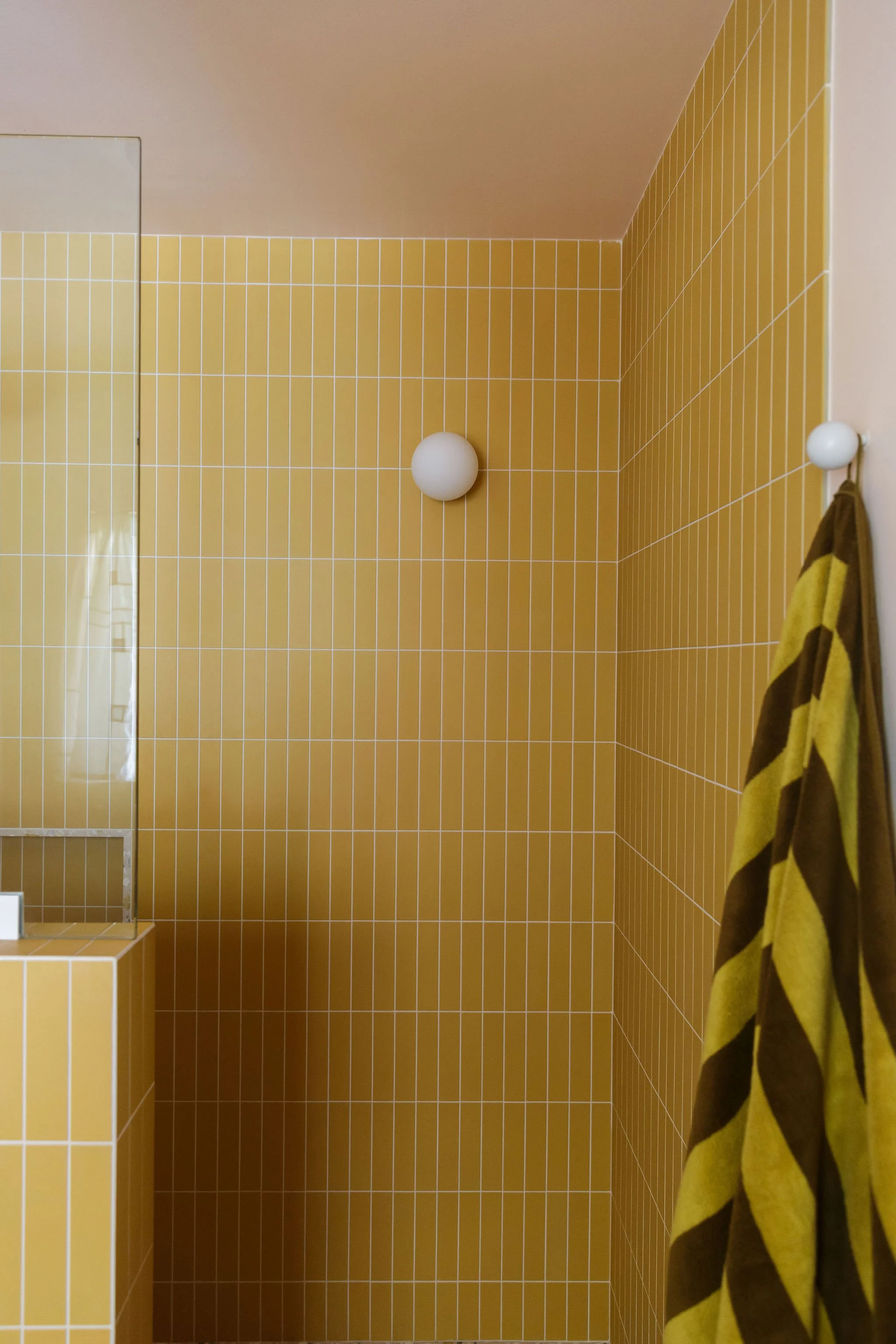Interior of a bathroom with yellow tiled walls, a white spherical wall light, a mirror, and a brown and yellow striped towel hanging on a white towel hook.