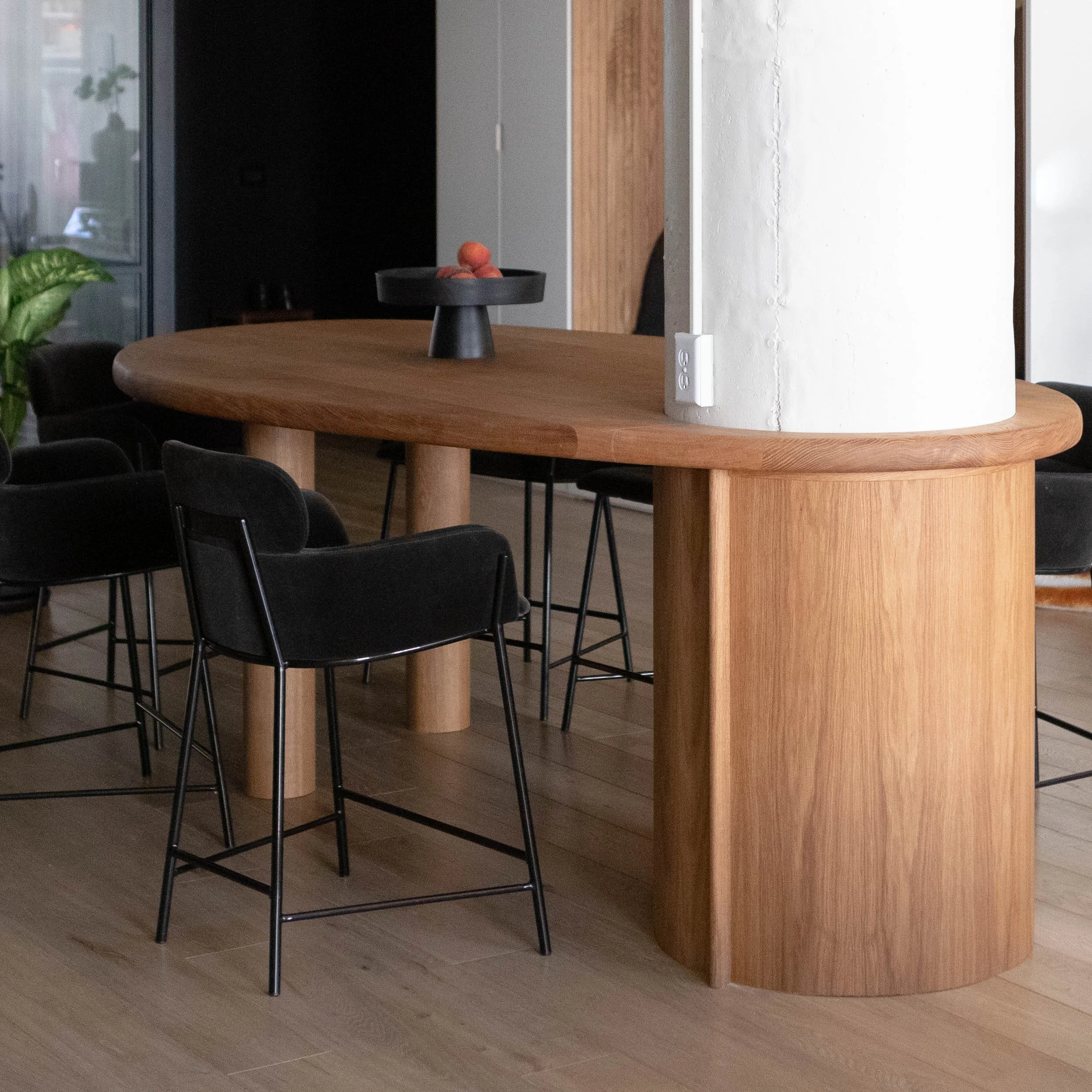 A wooden dining table with black chairs in a modern interior, with a black cake stand holding peaches on the table, and a white pillar with a small white device attached.