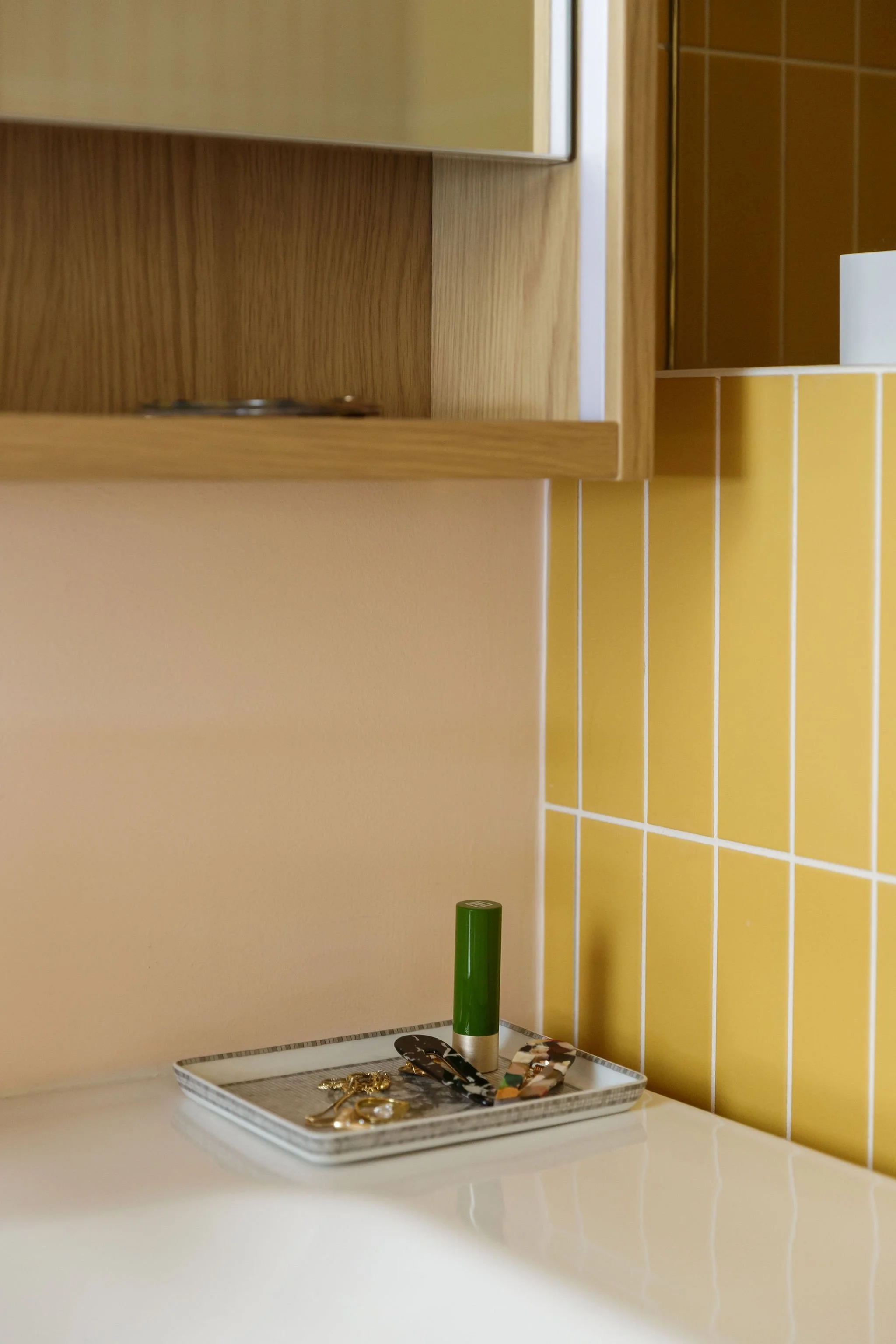 A bathroom vanity with a mirror, yellow tiled walls, and a tray holding jewelry and a green container.