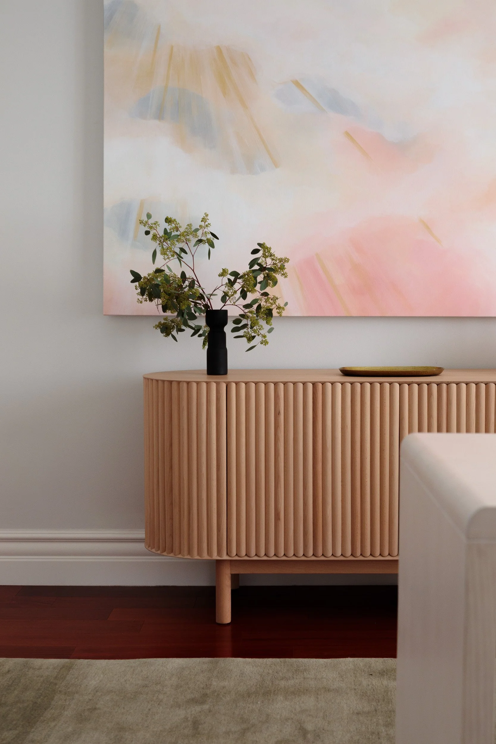A minimalist interior scene with a light wood textured sideboard, a black vase holding green leafy branches, and a gold tray, with a pastel abstract painting hanging above.