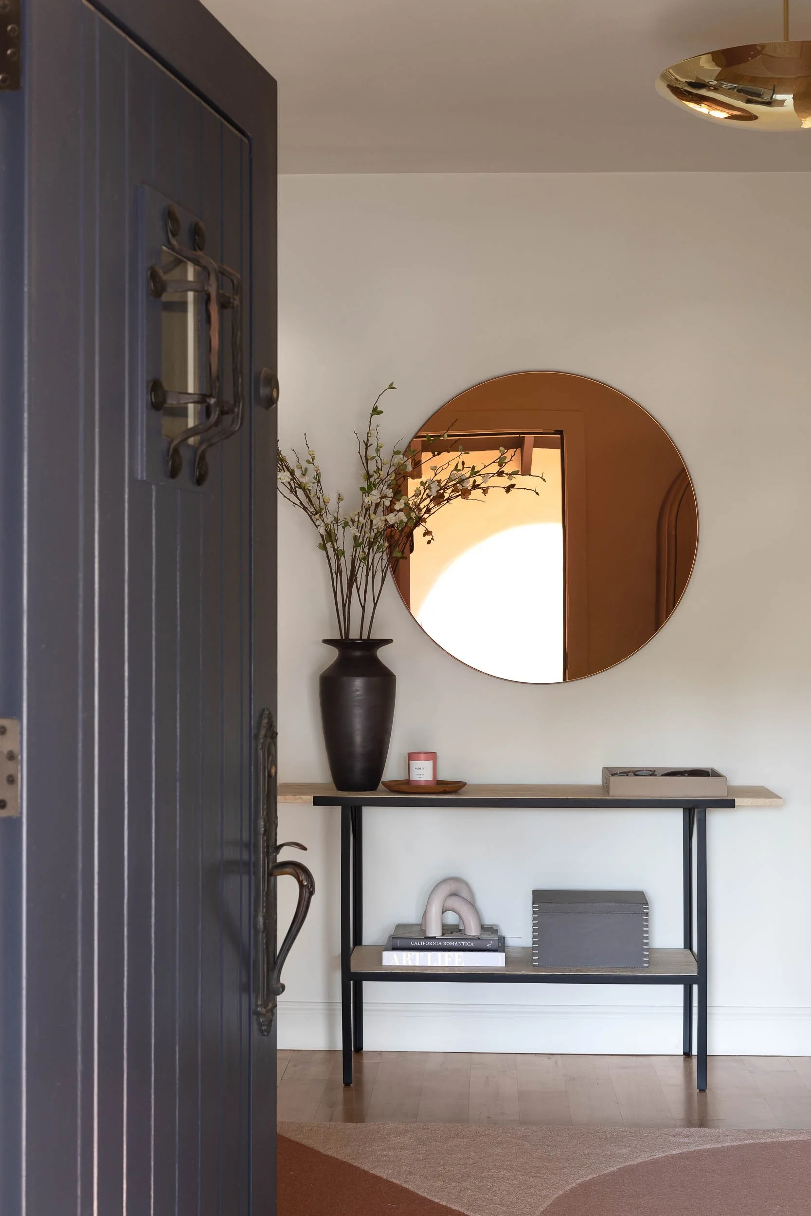 An entryway with a dark blue door opened to a hallway. Inside, there is a console table with a vase of branches with flowers, a candle, a small tray, and books underneath. A round mirror hangs on the wall above the table.