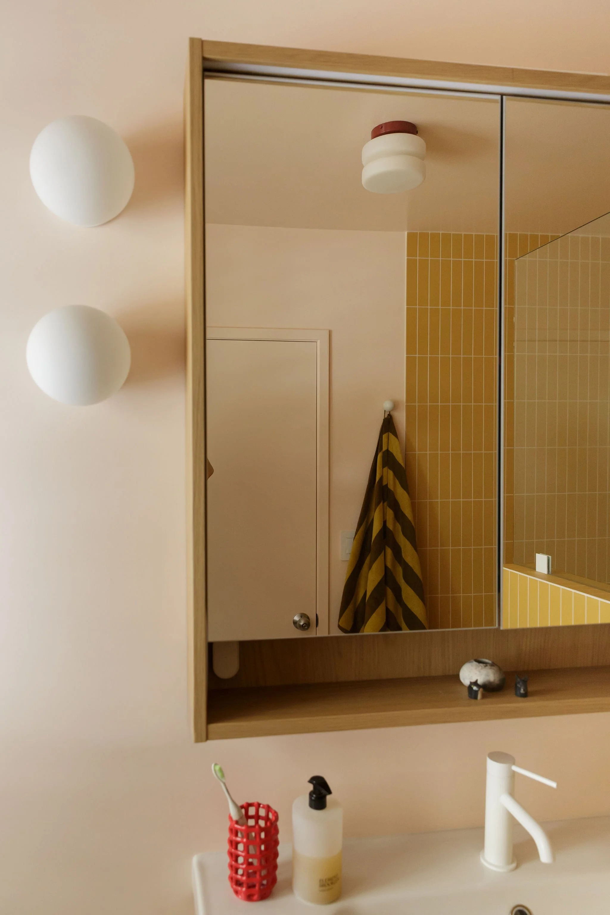 A bathroom mirror reflecting a wall with yellow tiles, a towel hanging, and a ceiling light. On the sink counter, there is a soap dispenser, a toothbrush in a red holder, and a candle. The wall beside the mirror has two round light fixtures.