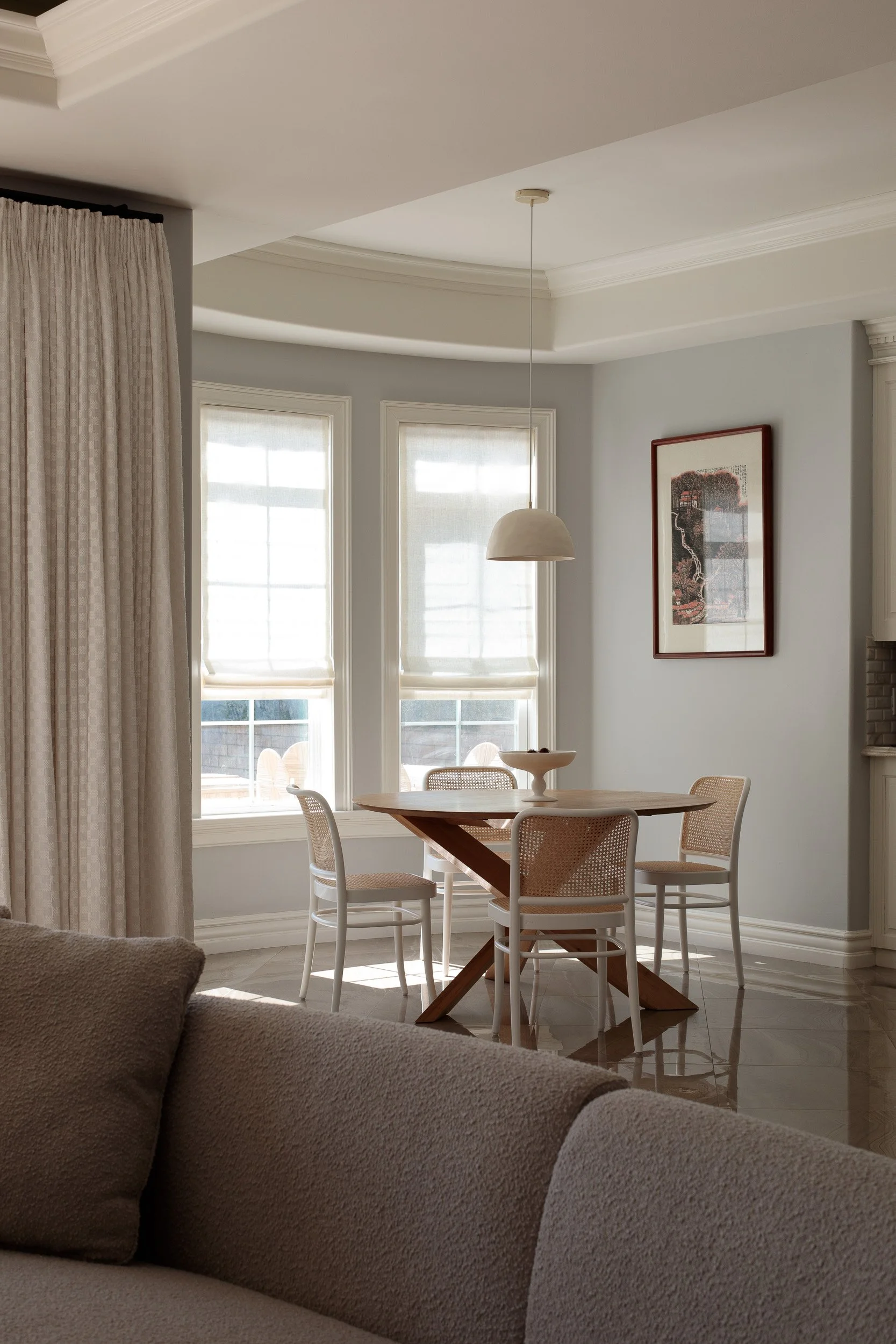 Dining area with a wooden round table, four white chairs, a pendant light, large windows with shades, and framed artwork on a light gray wall.