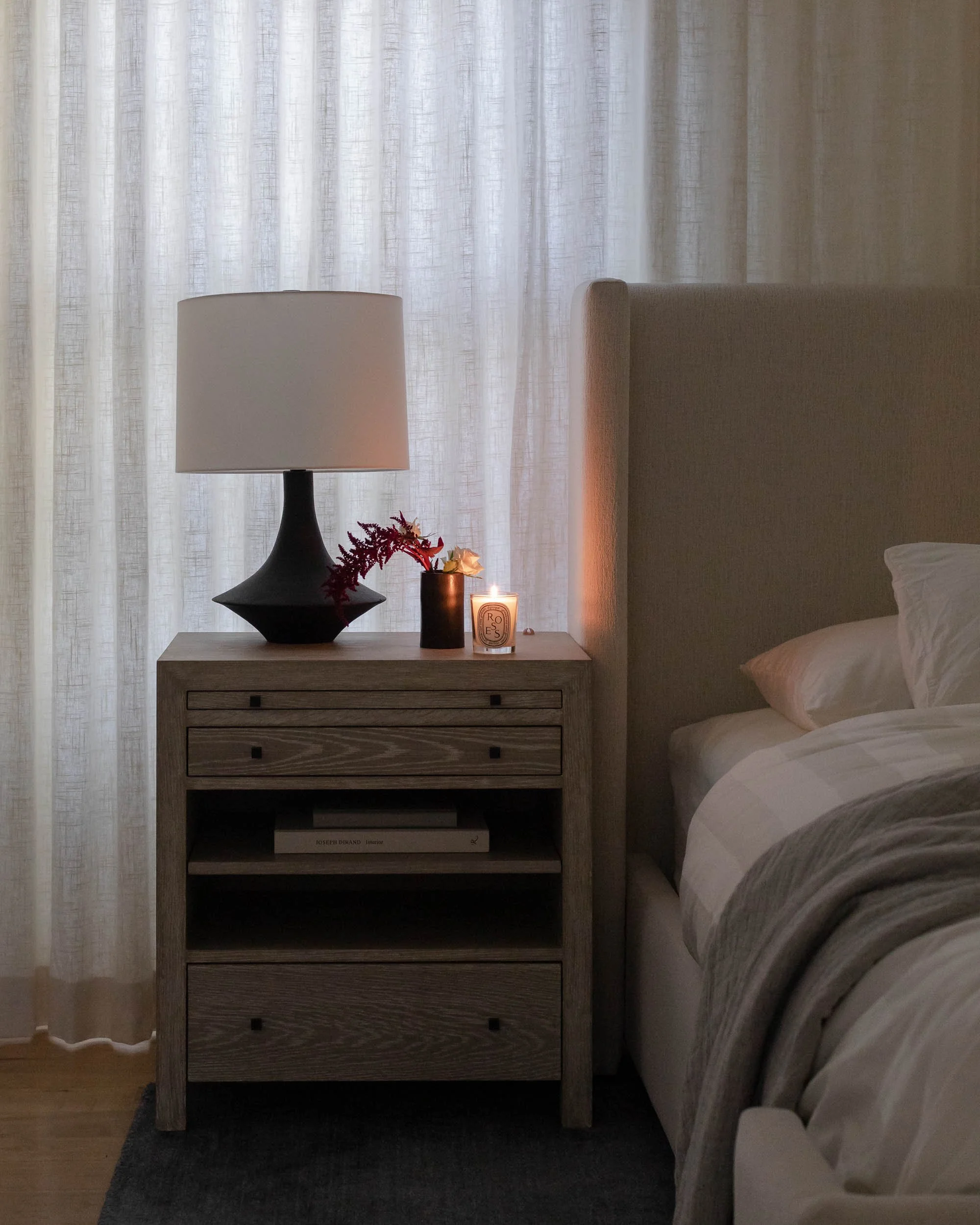 Bedroom nightstand with a lamp, a candle, and decorative flowers, by a bed with white and beige bedding, in front of sheer curtains.