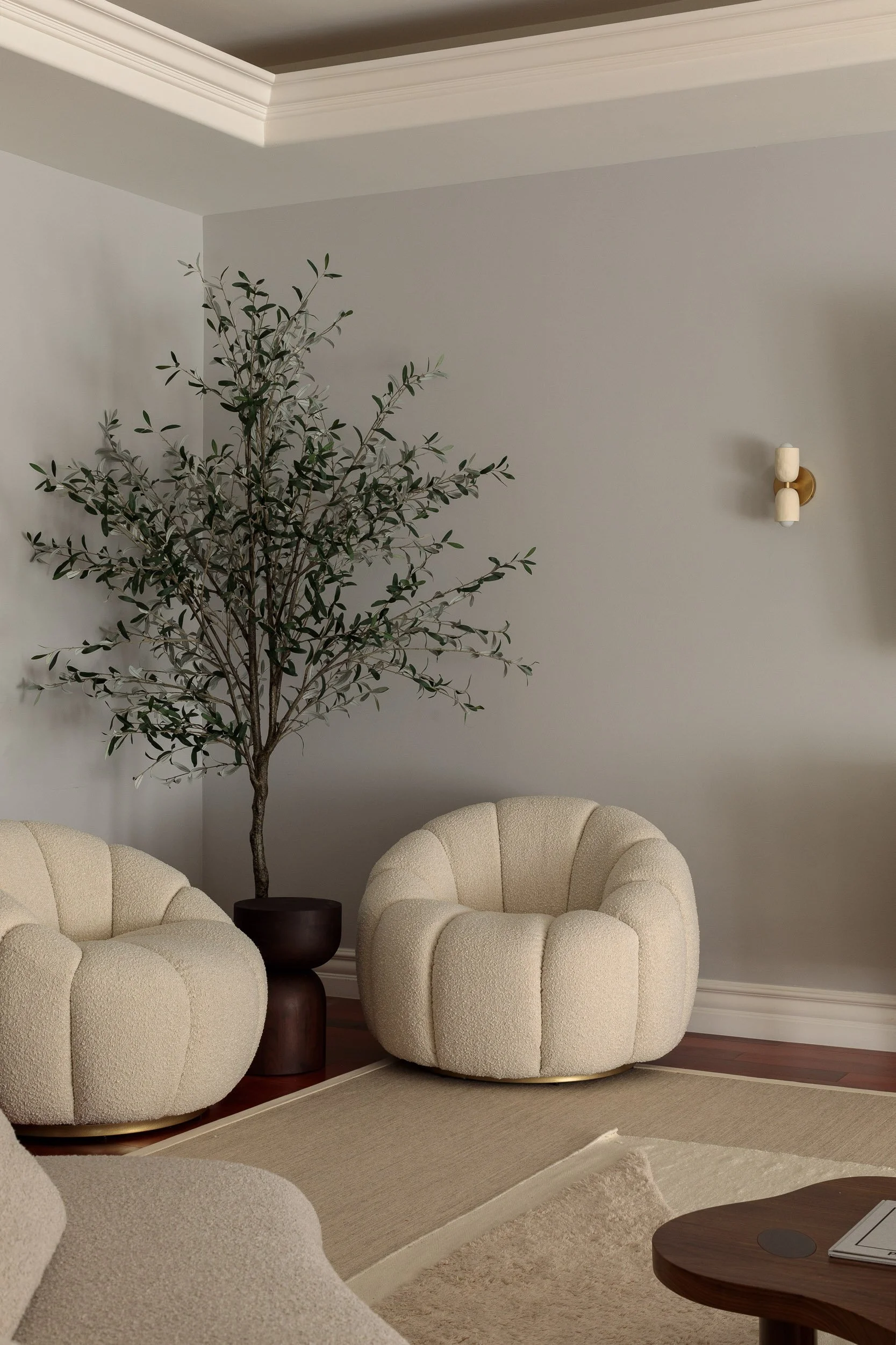 Living room with two plush cream-colored armchairs, a dark brown side table, and a large potted olive tree against a light gray wall. A beige rug and hardwood floor are visible.