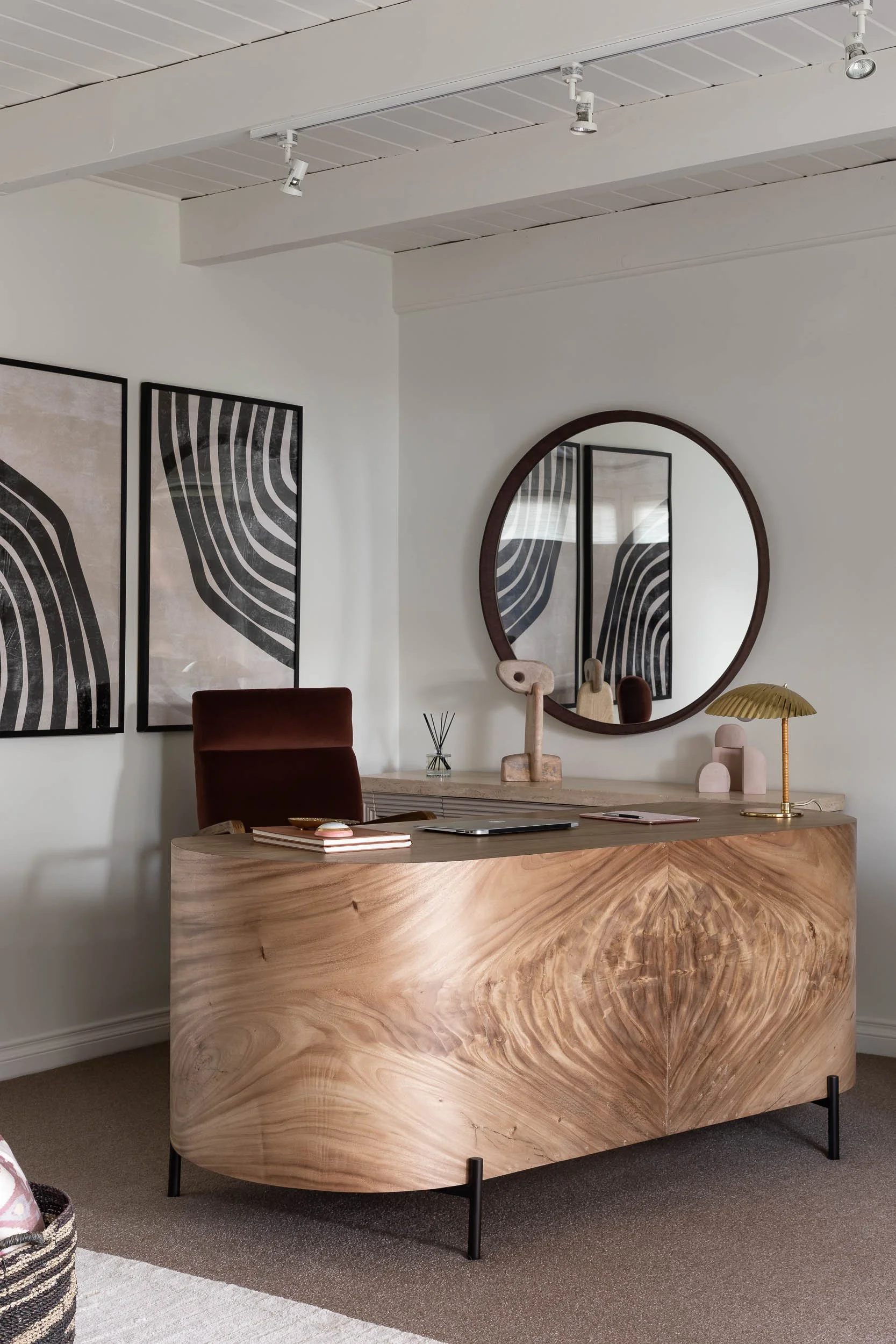 A modern office with a wooden desk, red velvet chair, round mirror, abstract art, and decorative objects.