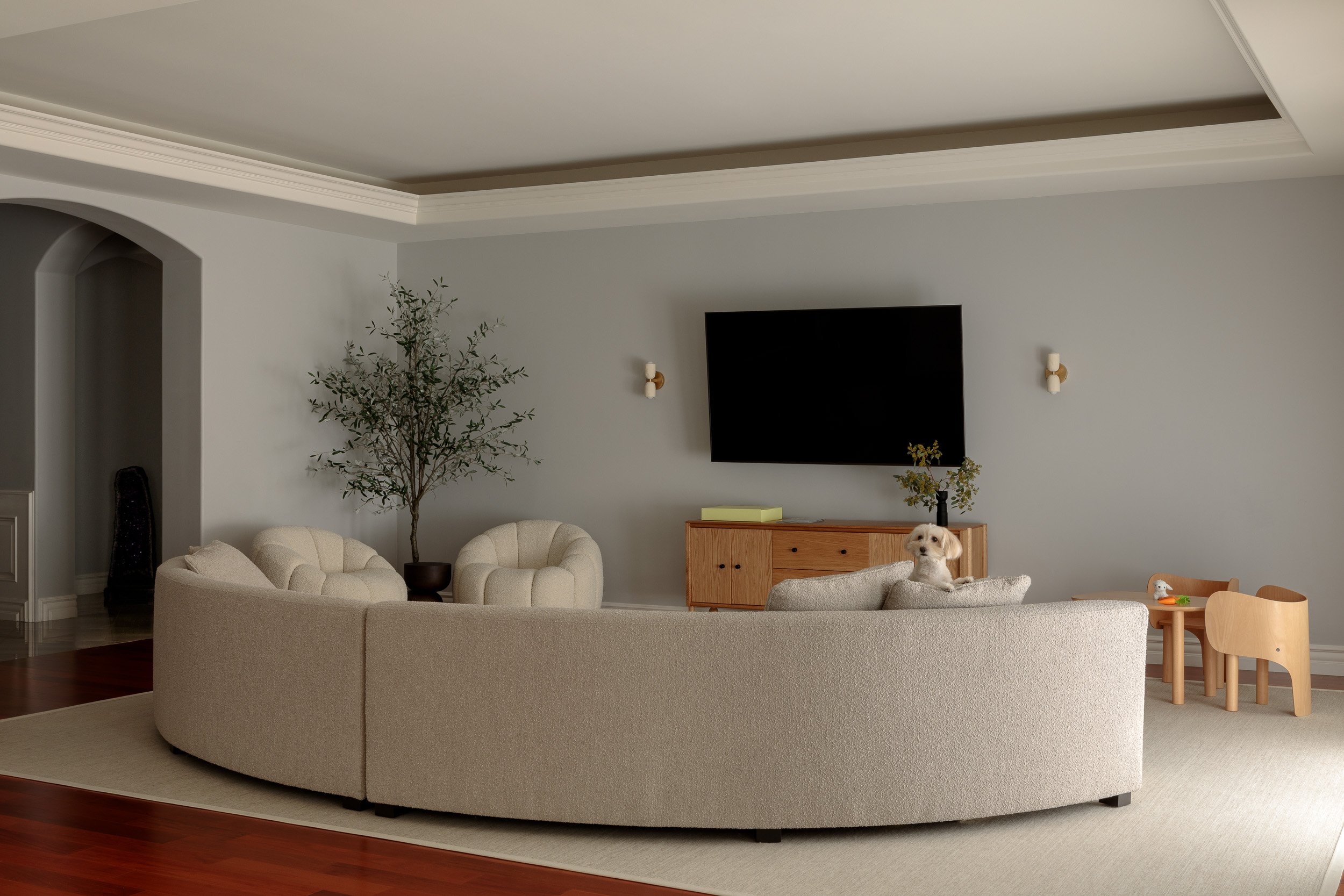 Living room with a beige curved sofa, a TV mounted on a gray wall, a small wooden table with a dog sitting on it, and children's toys on a small table. There is also a large potted plant.