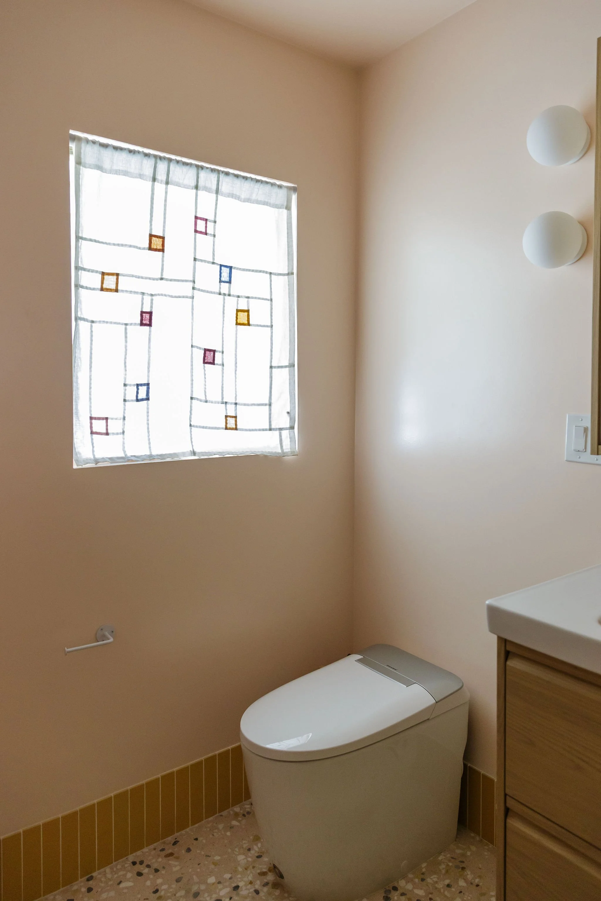 A bathroom with pink walls, a window with a colored geometric patterned curtain, a white modern toilet, a small wooden cabinet with drawers, a wall light fixture, and a terrazzo floor.