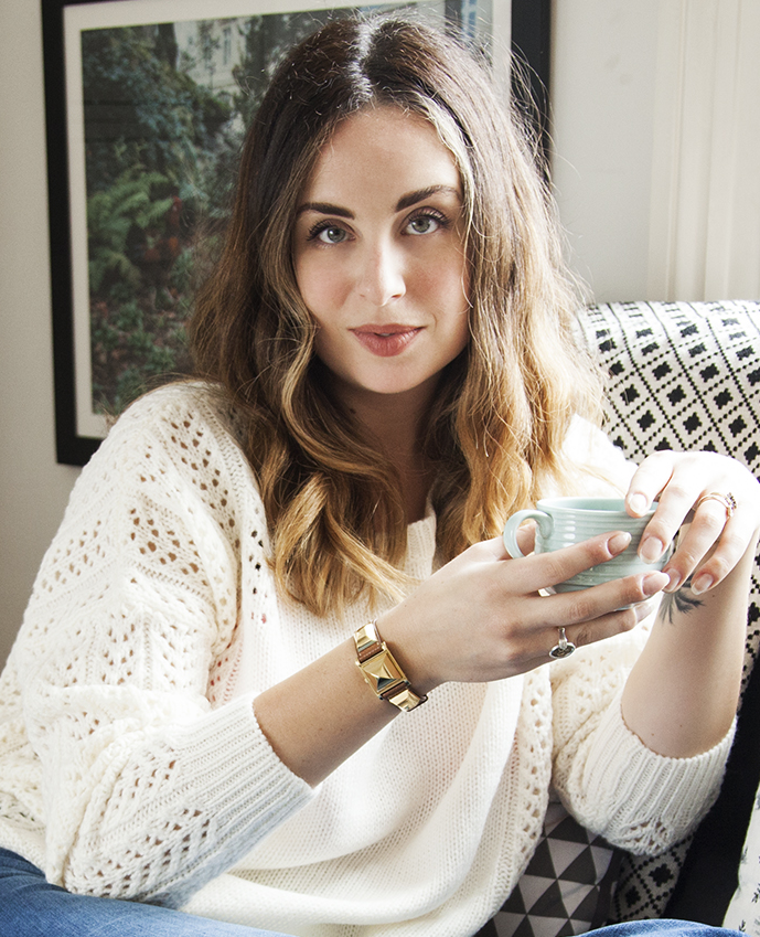 A woman with wavy, shoulder-length hair holding a teal ceramic mug, seated on a patterned cushion in a cozy, well-lit room.